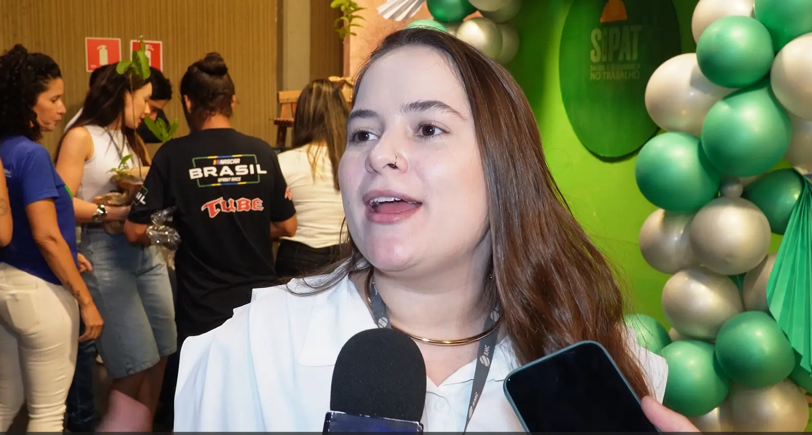 Close de uma mulher de cabelos castanhos falando ao microfone. Ela usa um colar dourado e crachá. Ao fundo, balões coloridos e outras pessoas de costas.