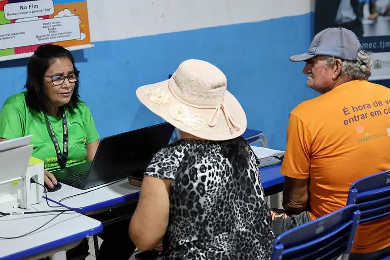 Foto horizontal colorida, em plano aberto, que mostra a senhora Rita Maria sentada, sendo atendida por servidora do CRAS, em uma sala de aula, onde ocorreu o mutirão Justiça em Ação.