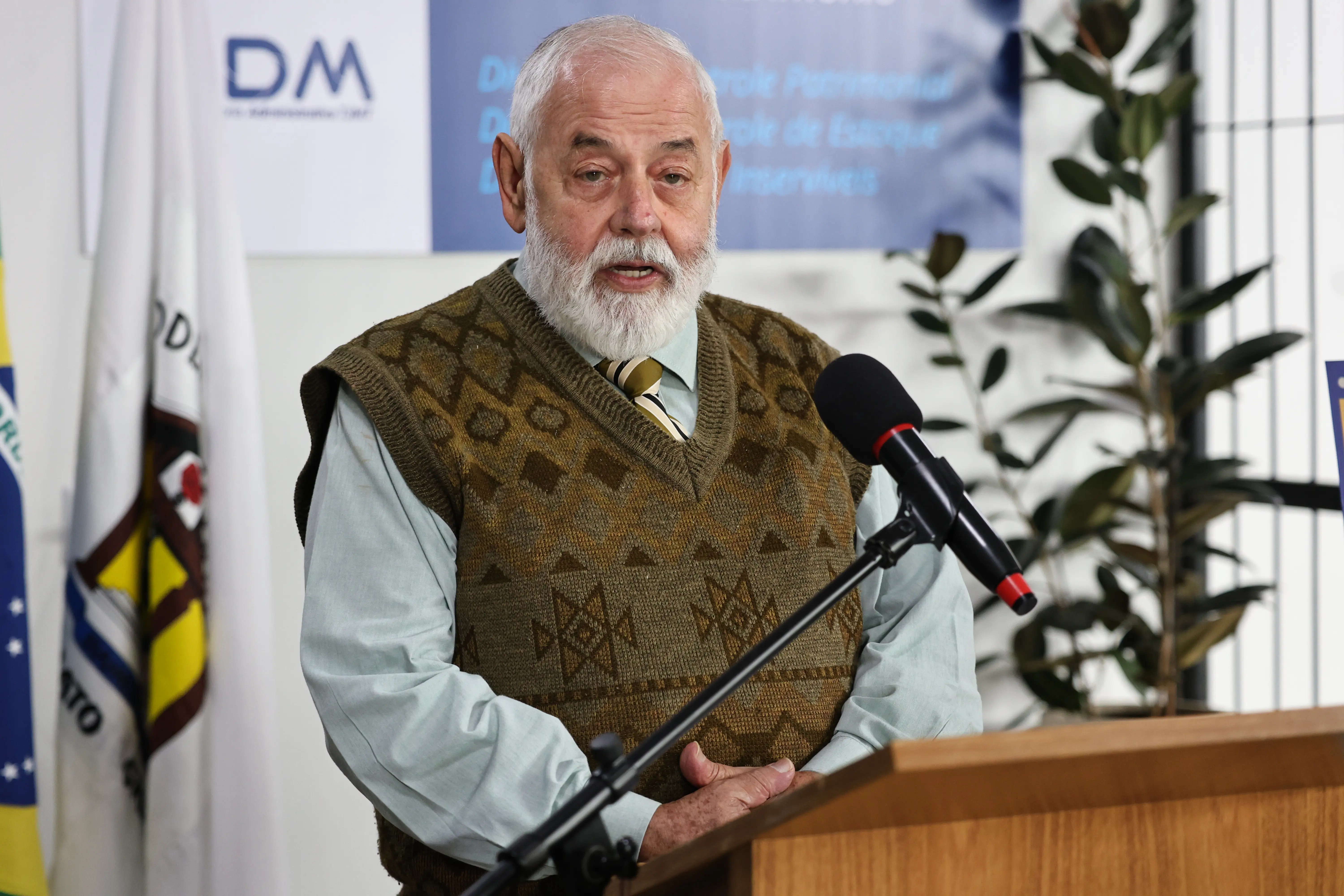 Homem idoso de pele clara e barba branca fala ao microfone em um púlpito de madeira. Ele veste camisa clara, gravata listrada e um colete de lã marrom com estampas geométricas.