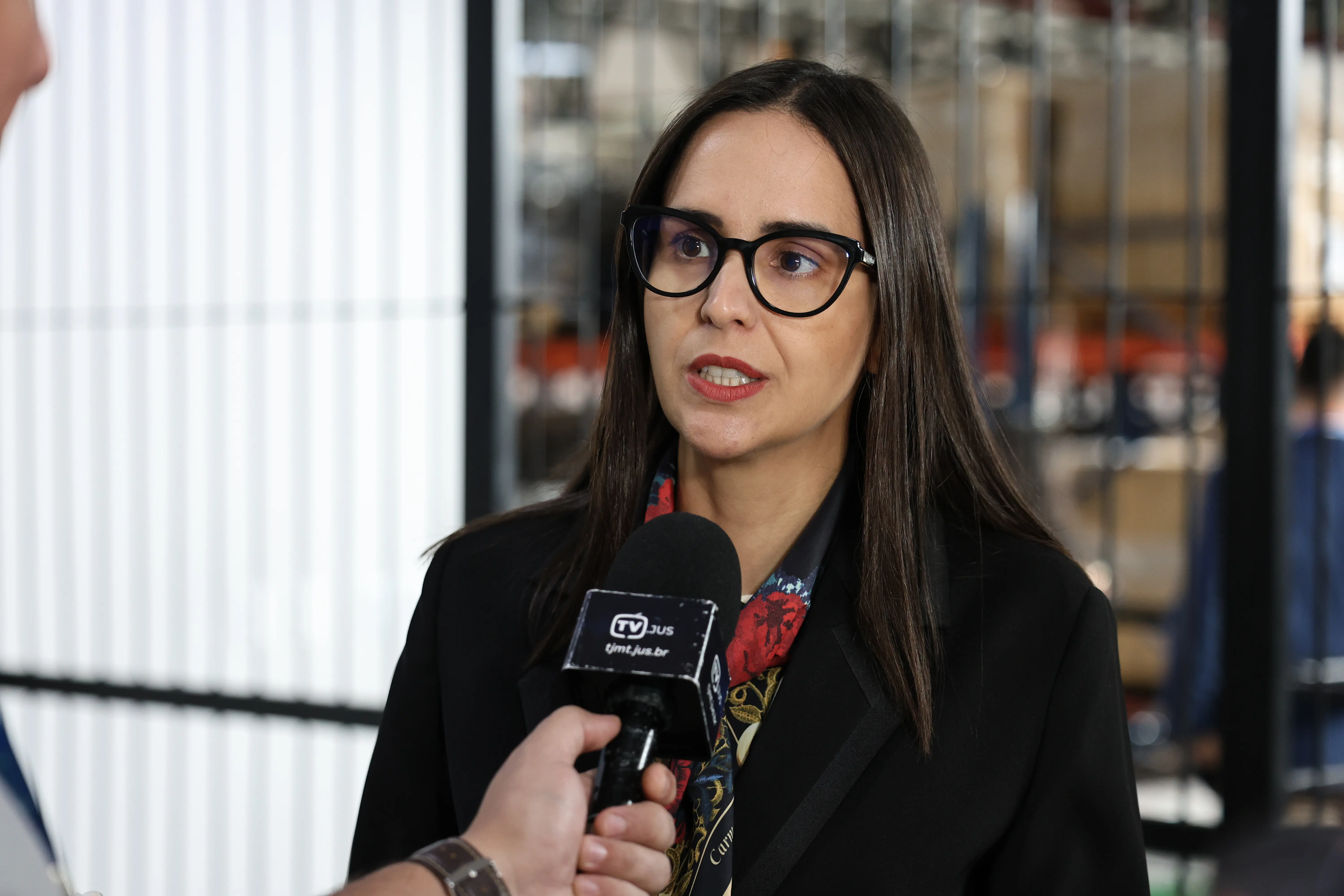Mulher de pele clara, cabelos longos e escuros, usando óculos de armação preta e blazer preto, fala diretamente para um microfone de reportagem com o logotipo da TV Jus.