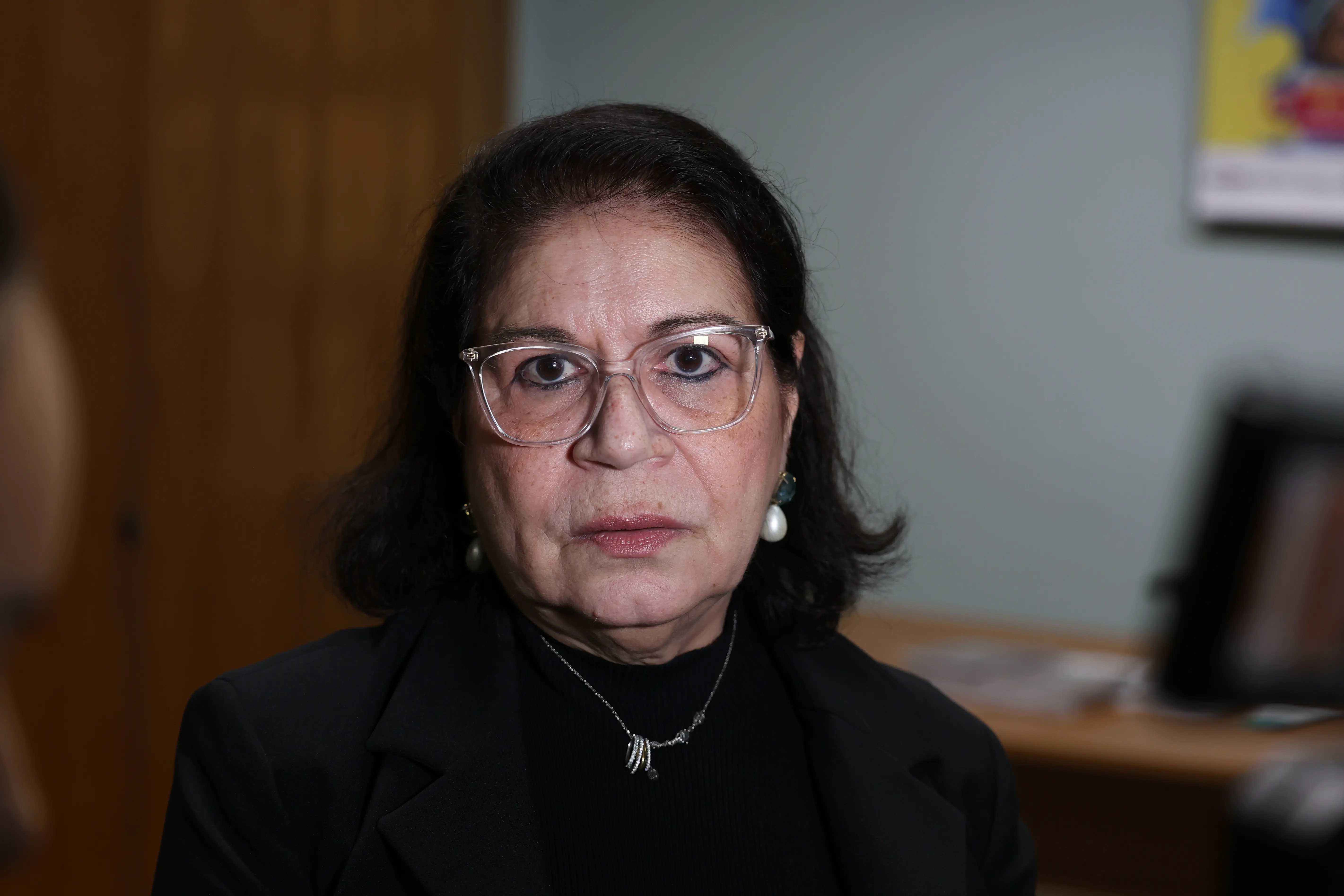 Retrato frontal de uma mulher. Ela usa óculos de armação transparente, brincos de pérola, colar delicado e blazer preto sobre blusa de gola alta escura.