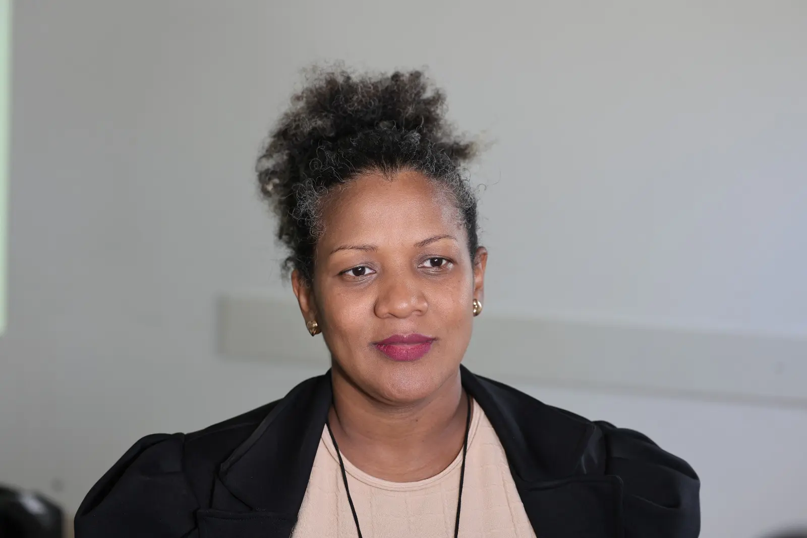 A imagem mostra a secretária adjunta de Assistência Social, Vilmara Vidics. Ela aparece em primeiro plano, olhando para a câmera, usando blazer preto sobre blusa em tom claro. Ao fundo, o ambiente da palestra aparece desfocado.