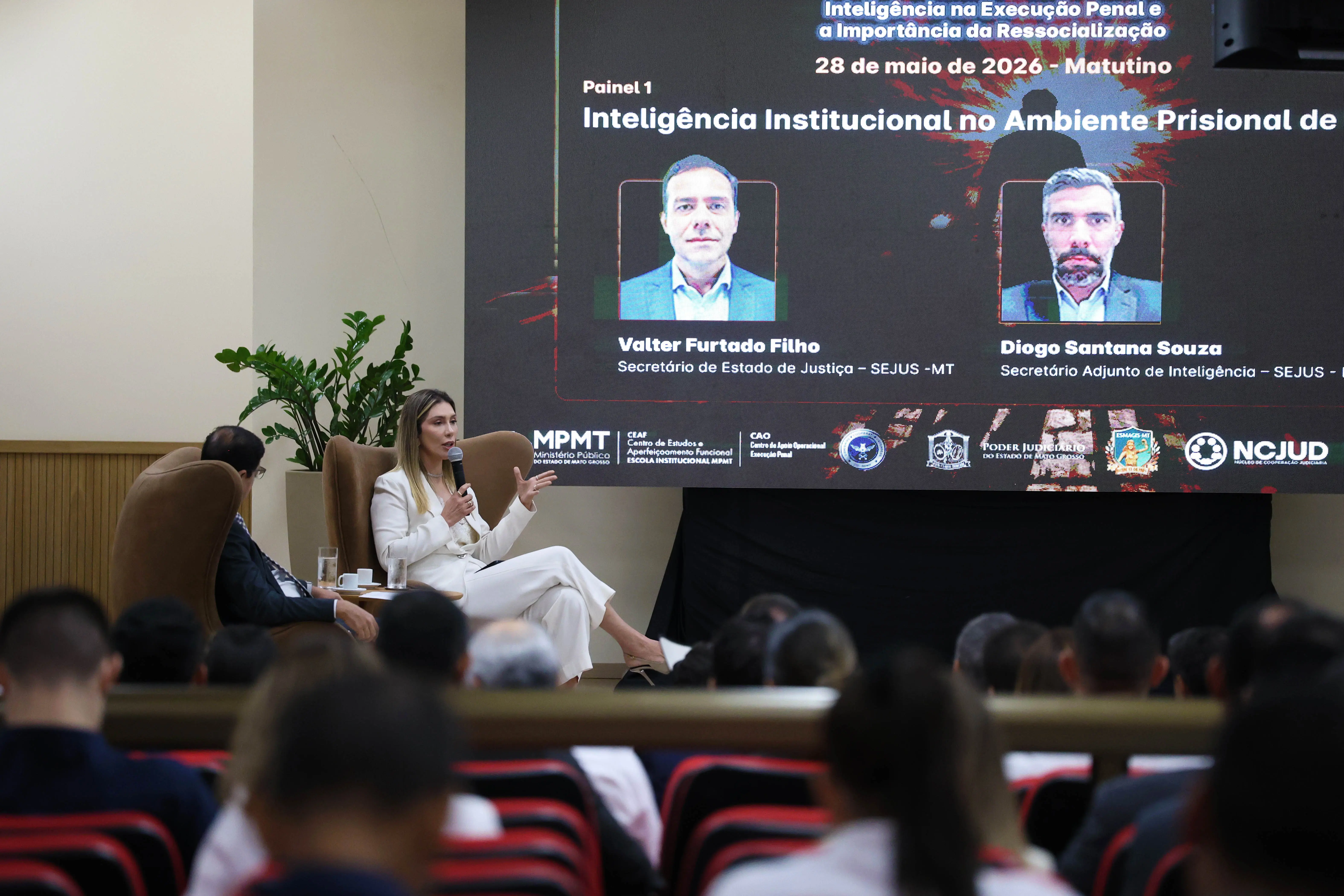 Mulher loira de terno branco fala ao microfone sentada em poltrona marrom no palco. Ao fundo, um grande telão exibe fotos e nomes de palestrantes. Em primeiro plano, a nuca da plateia sentada.