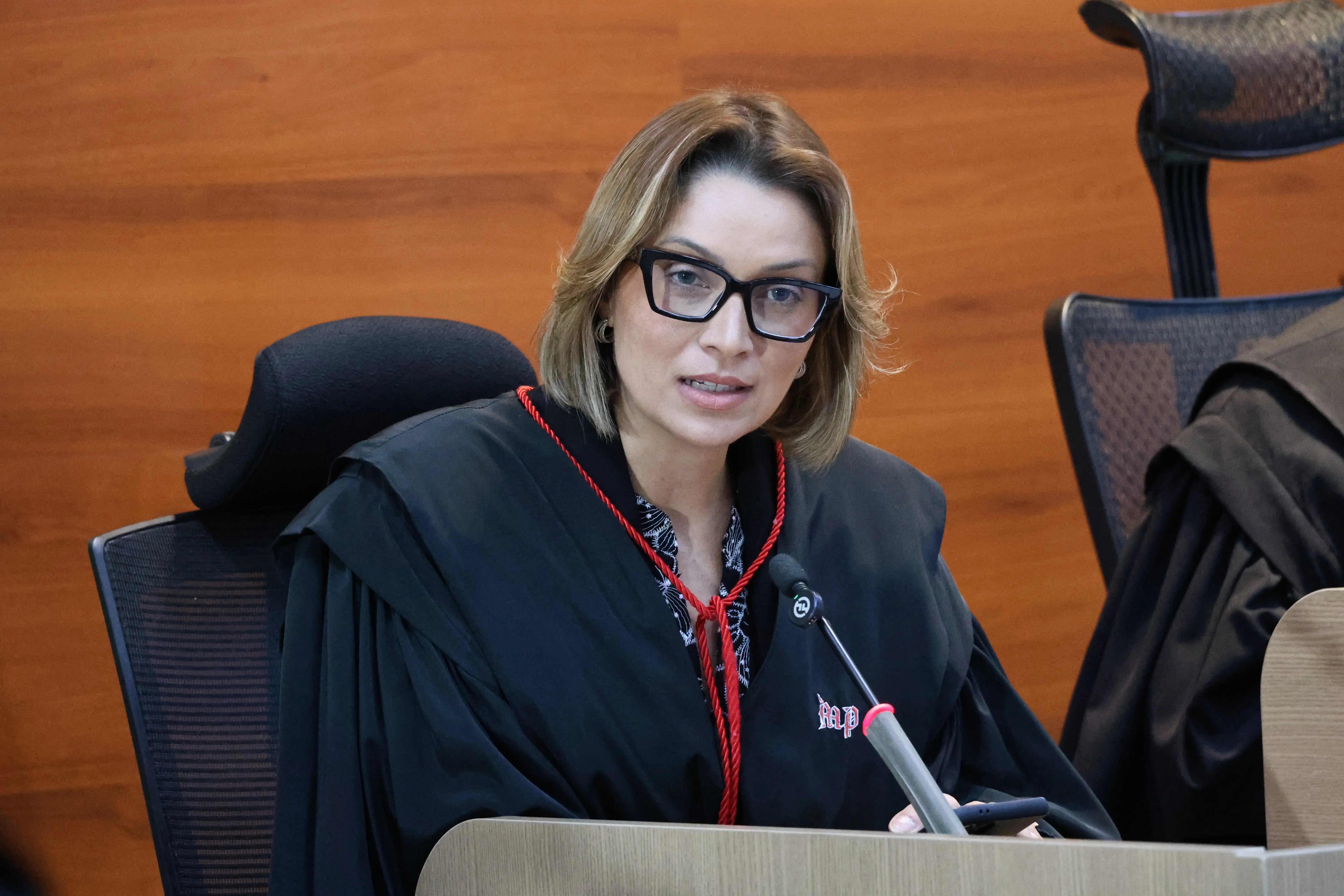 Mulher branca de cabelos loiros curtos e óculos fala próxima a um microfone de bancada no plenário. Veste veste preta com cordão vermelho e a sigla MP. Ao fundo, painel de madeira desfocado.