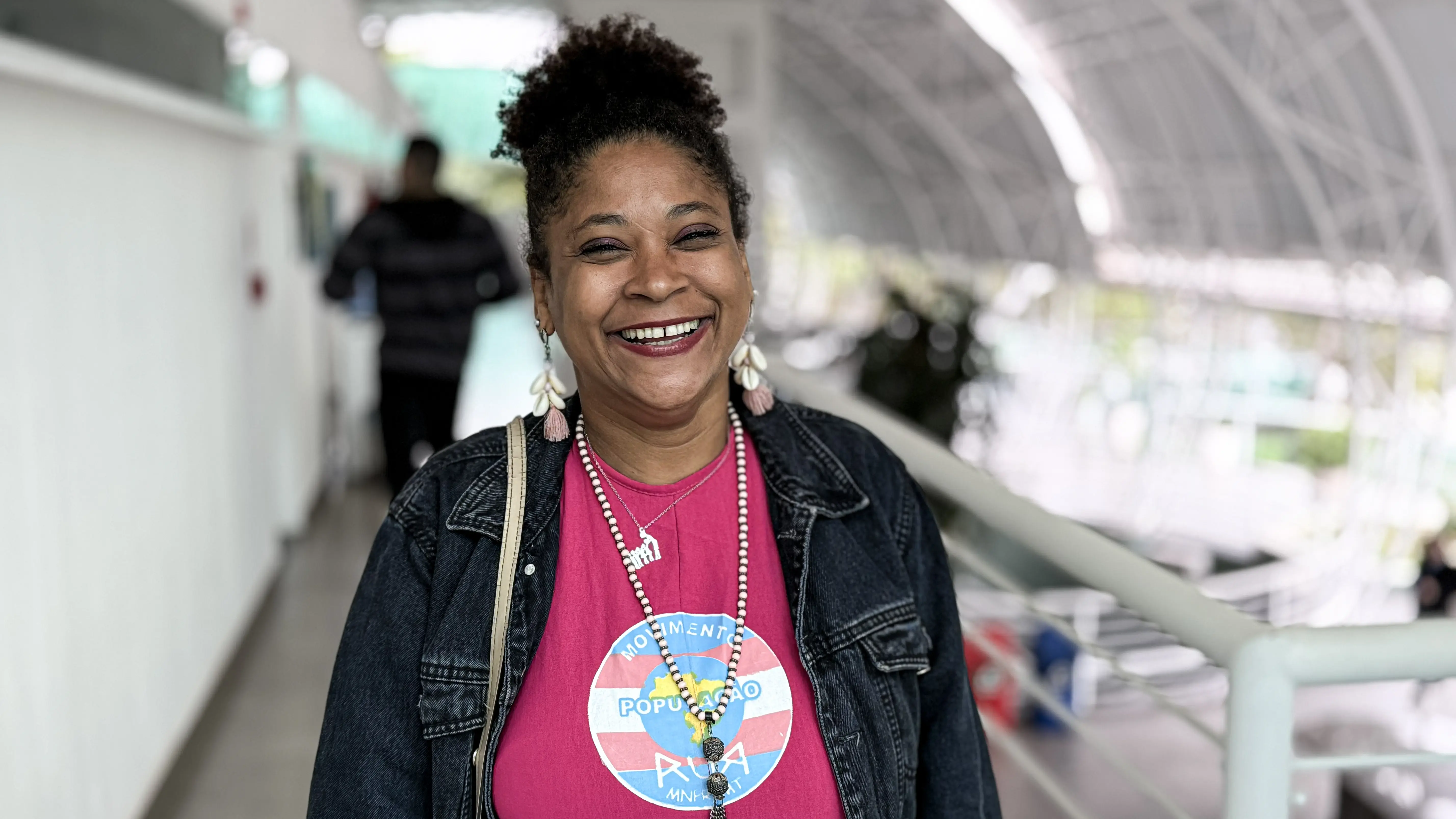 Mulher negra de cabelos crespos sorri amplamente. Veste camiseta rosa com logotipo do Movimento População de Rua e jaqueta jeans.