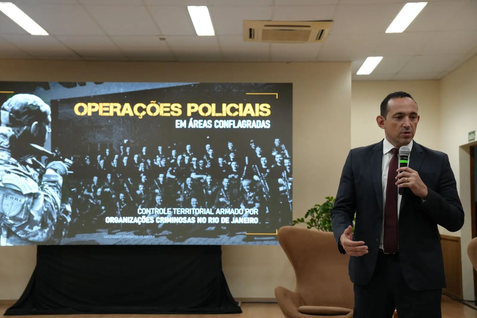 Foto horizontal que mostra o delegado de polícia civil do Rio de Janeiro, Fabrício Oliveira, proferindo palestra. Ele é um homem branco, de cabelos escuros, usando camisa branca, terno cinza e gravata vermelha. Atrás dele, há um telão com imagem de policiais e o tema Operações Policiais. 
