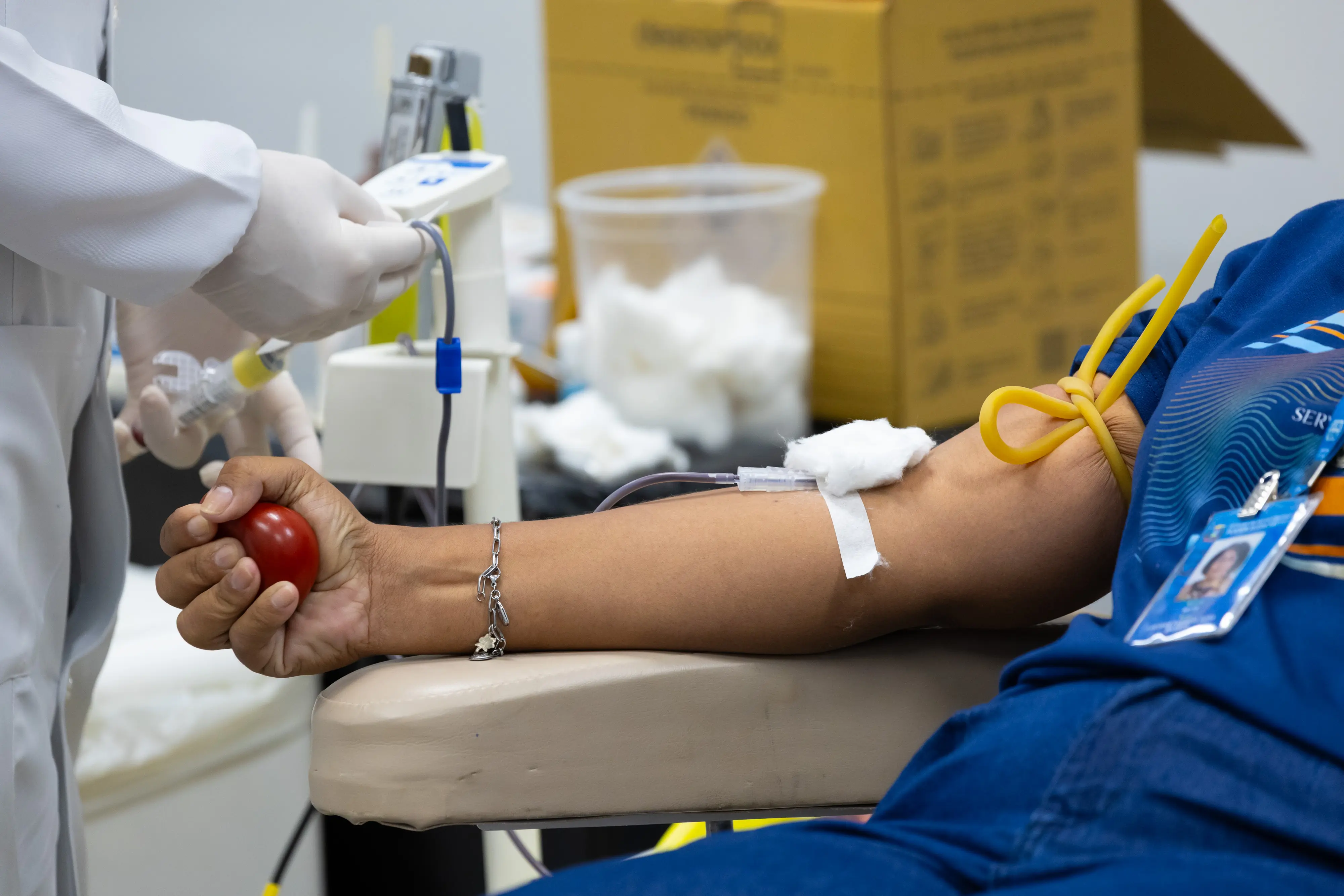 Close no braço de um doador apertando uma bolinha vermelha enquanto uma profissional de saúde, de luvas brancas, prepara a coleta de sangue.