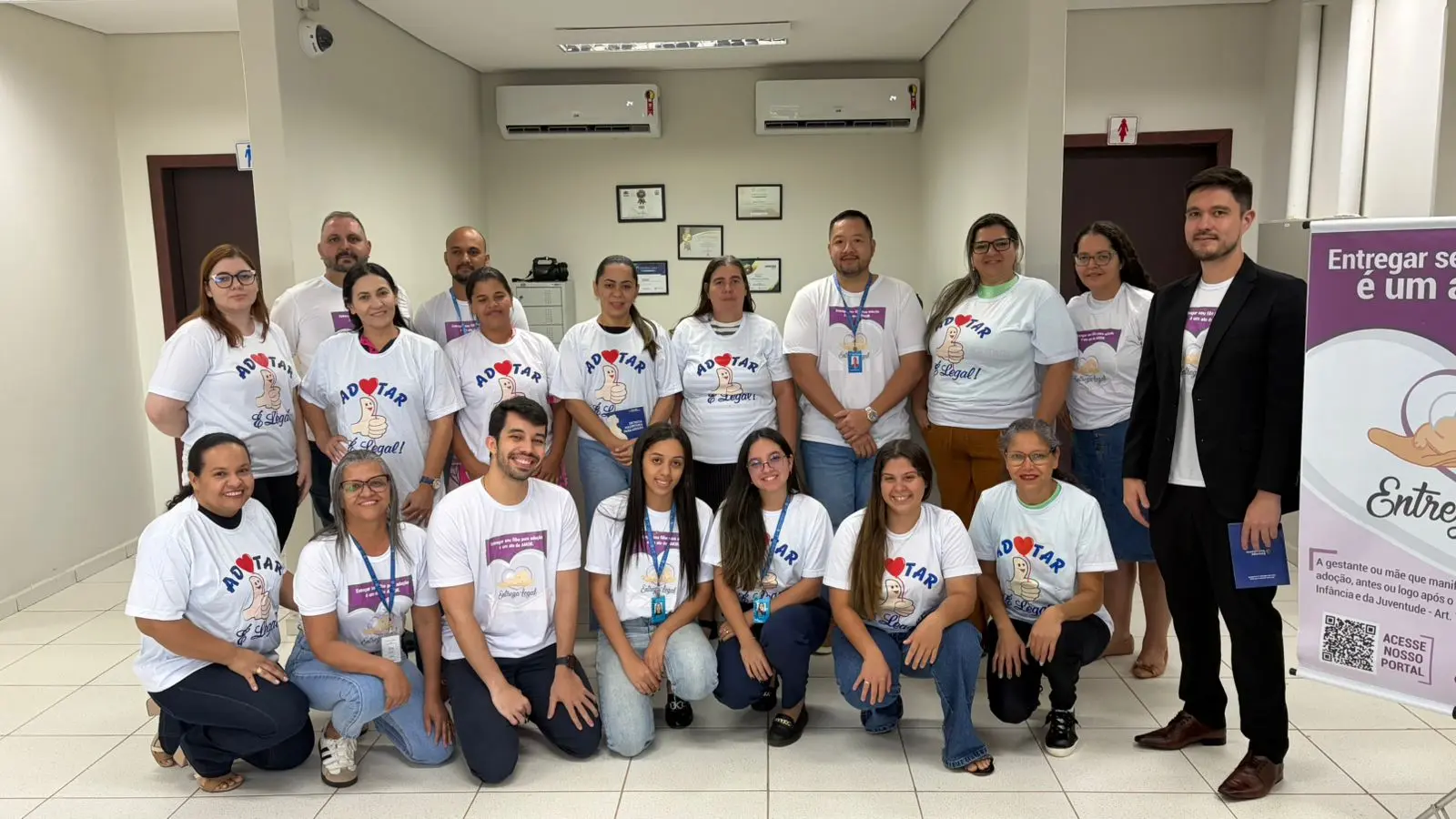 Foto horizontal colorida em plano aberto que mostra 18 pessoas posando para a foto, sorrindo. Todos usam camiseta da campanha Adotar é Legal. 
