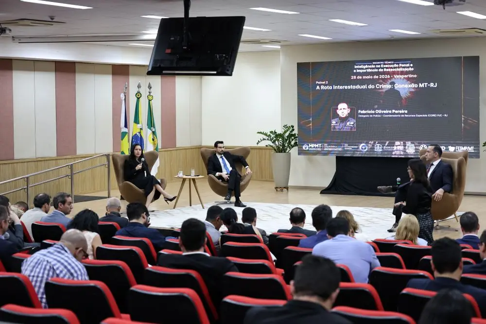 Foto horizontal em plano aberto que mostra um auditório visto do fundo, com pessoas sentadas na plateia e, no palco, dois homens e uma mulher sentados em poltronas. No telão, aparece o tema da palestra e foto do palestrante.