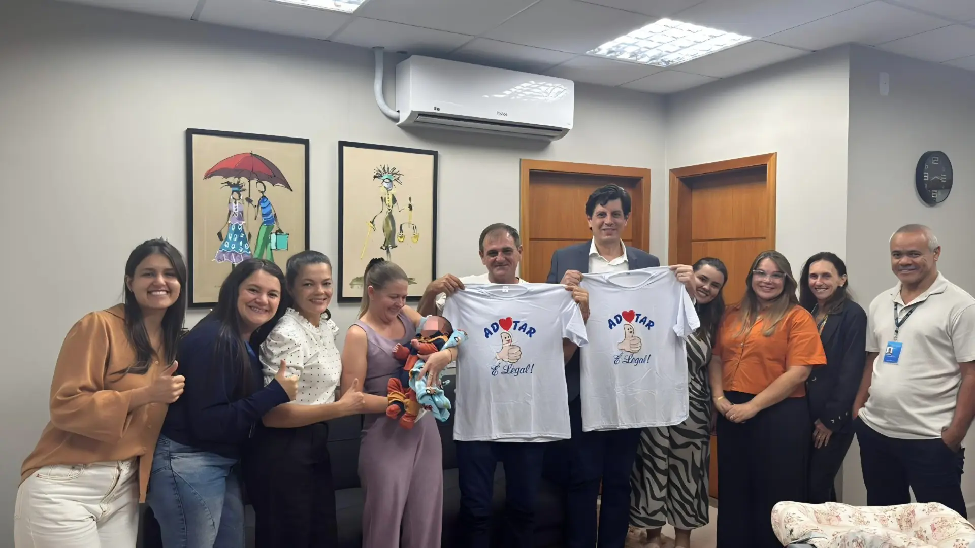 A equipe da 2ª Vara Cível está perfilada em pé, uma mulher segura um bebê no colo, com o rosto preservado digitalmente para garantir sua identidade. Ao lado dela, dois homens exibem camisetas brancas com a frase 