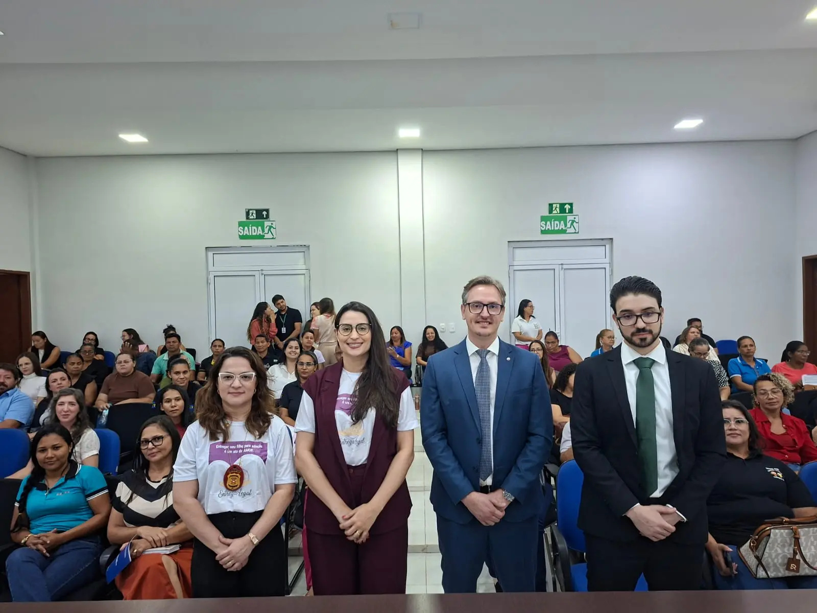 Fotografia mostra o ambiente interno do tribunal do júri, em primeiro plano estão os palestrantes, duas mulheres e dois homens, perfilados lado a lado. Ao fundo os participantes do evento, que estão sentados.
