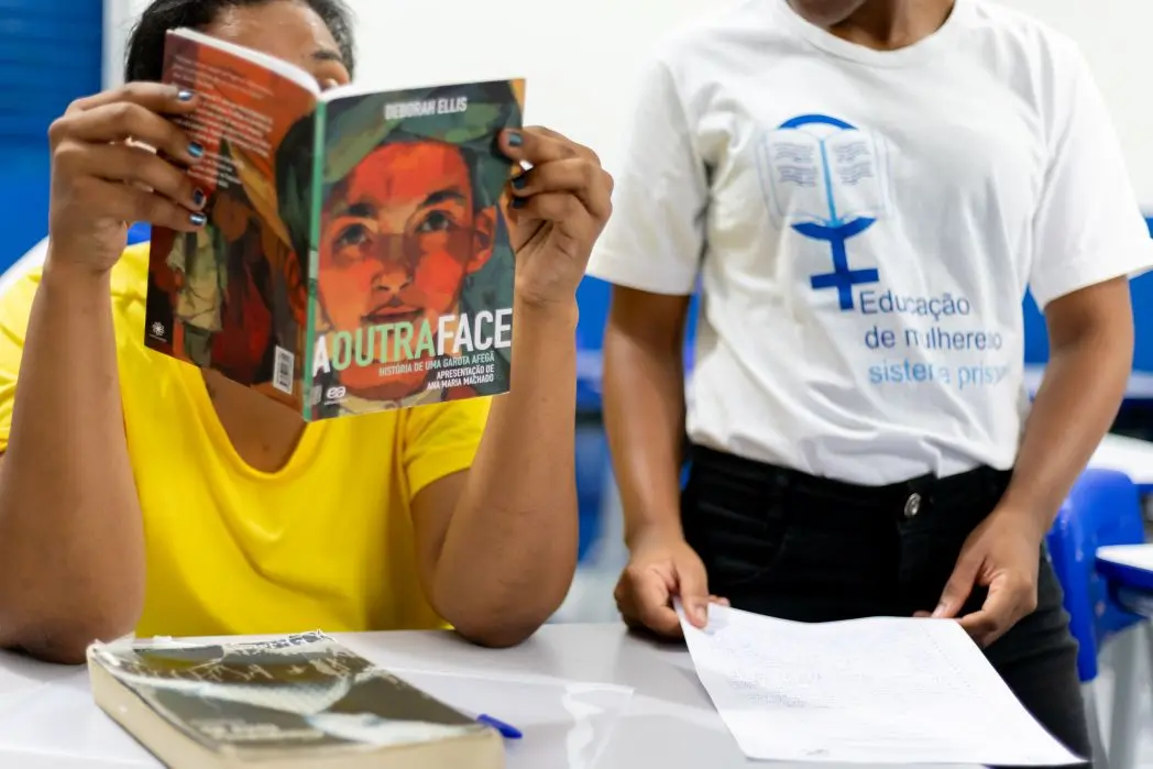 Foto horizontal que mostra uma mulher privada de liberdade em sala de aula, sentada, lendo o livro 