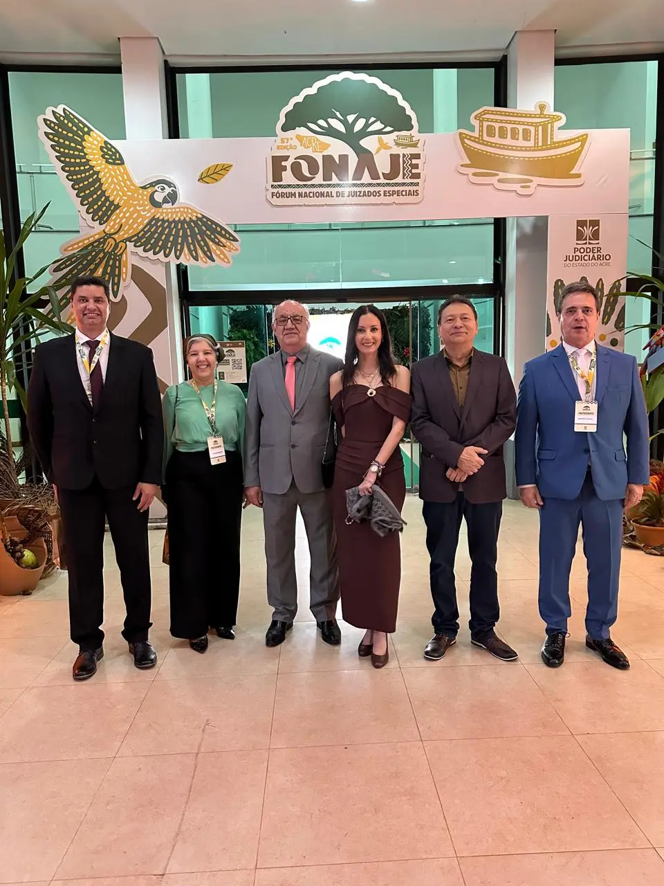 Foto vertical que mostra quatro magistrados e duas juízas de Mato Grosso em pé, posando para a foto. Eles estão em um espaço de convenções. Ao fundo, há um banner do Fonaje.