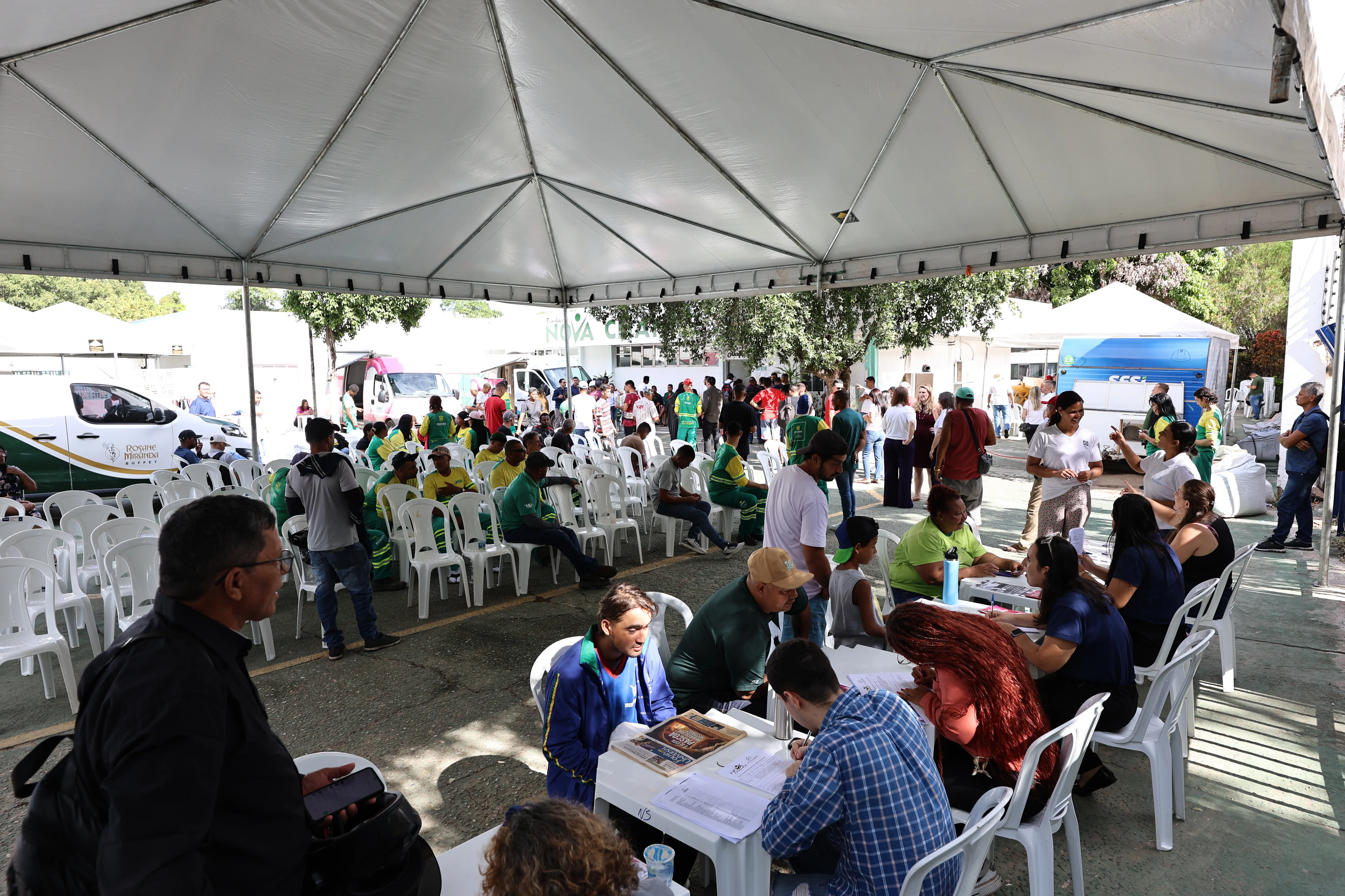 Vista ampla sob tenda branca com várias cadeiras ocupadas por pessoas em uniformes verdes e amarelos. Há mesas de atendimento e veículos brancos estacionados ao fundo.