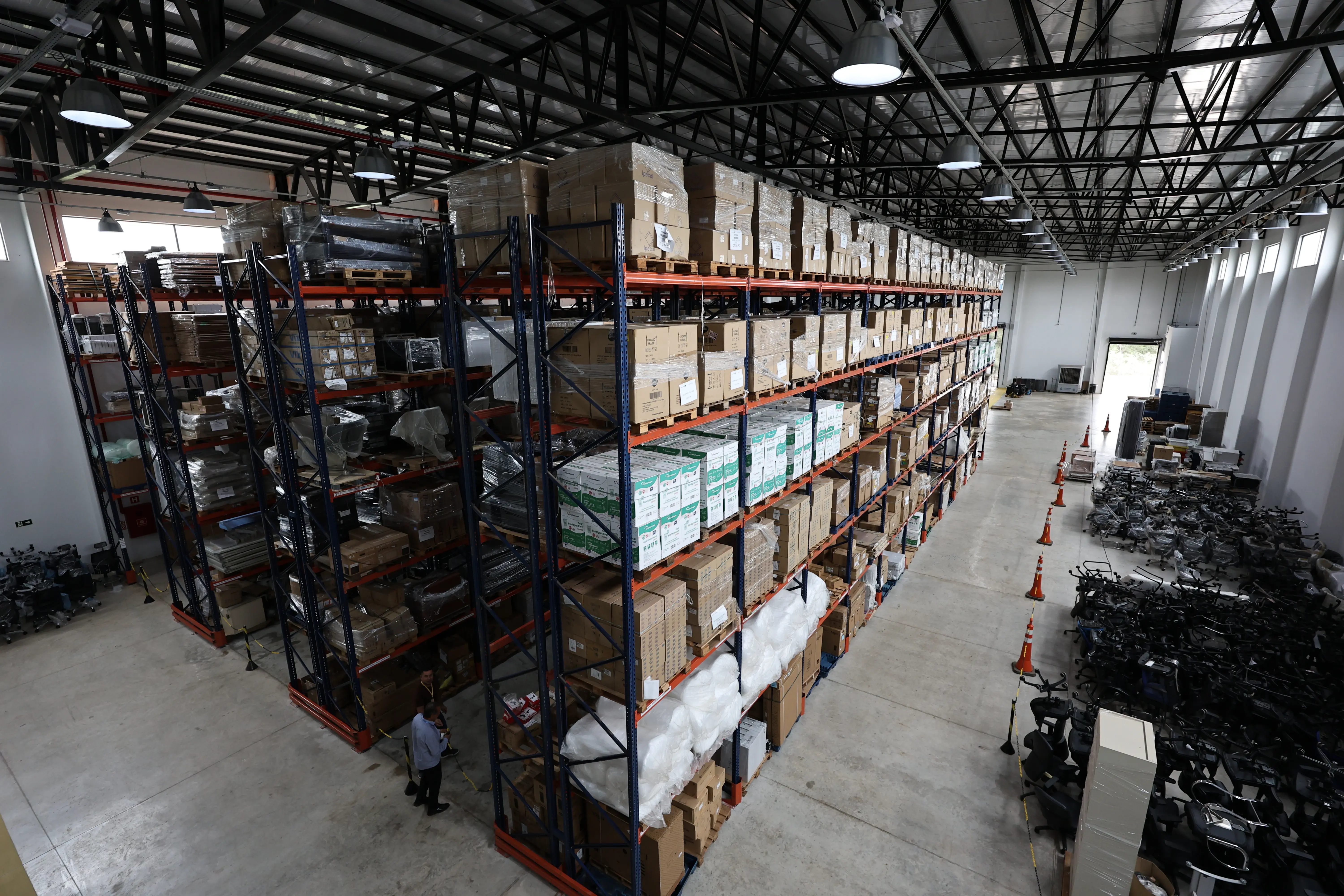 Visão aérea do interior de um grande galpão logístico, mostrando longas e altas estruturas metálicas repletas de caixas de papelão organizadas em paletes, com duas pessoas andando no corredor.