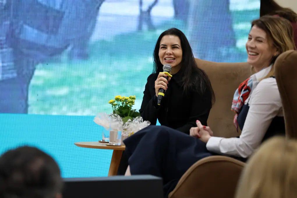Foto em plano médio de duas mulheres sentadas em poltronas durante um evento. À esquerda, uma mulher de cabelos pretos sorri e fala ao microfone. À direita, outra mulher ri, de perfil. Ao fundo, um grande painel de LED exibe uma imagem desfocada.
