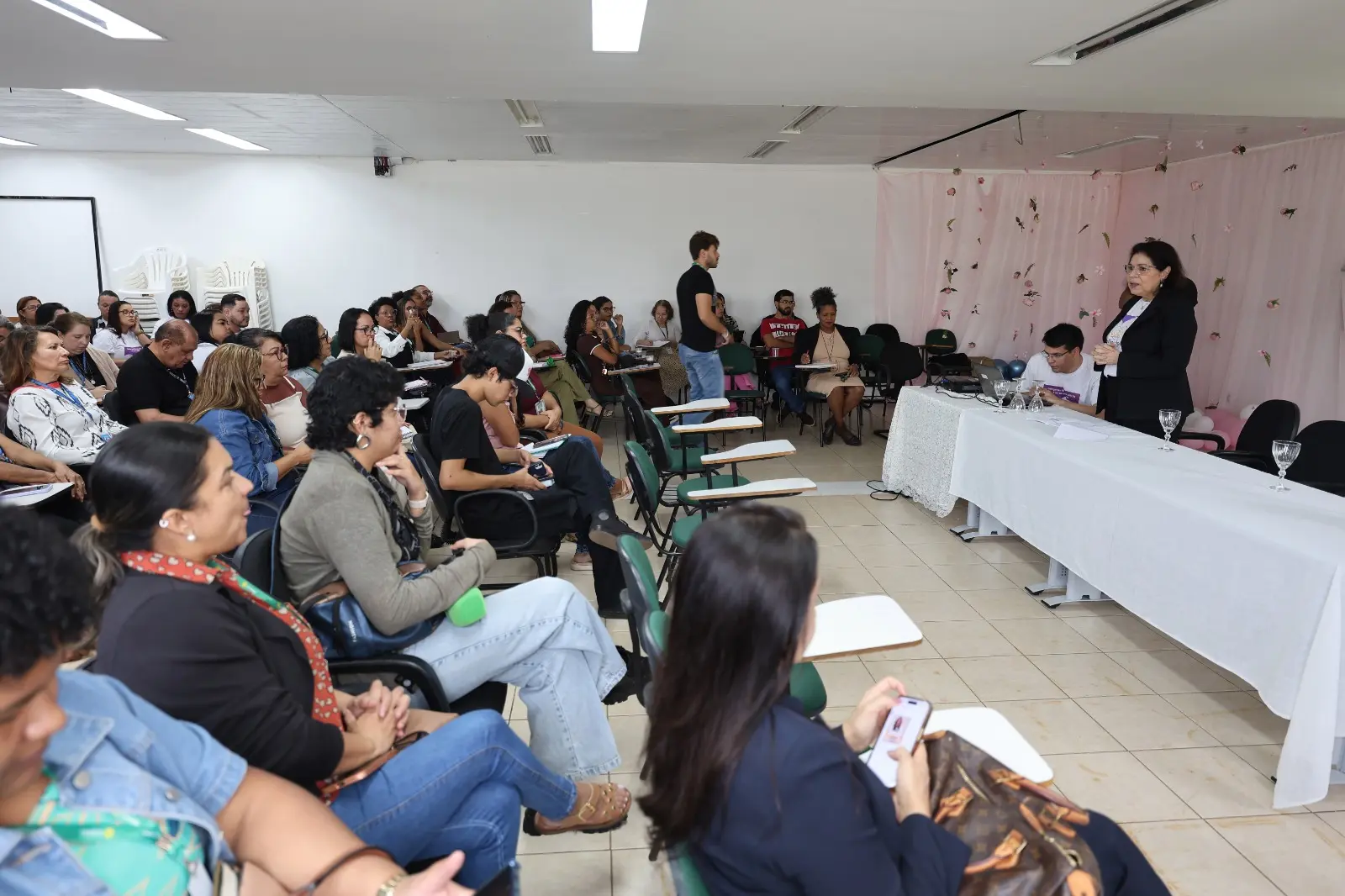 A juíza da 1ª Vara Especializada da Infância e Juventude de Cuiabá, Gleide Bispo Santos, aparece em pé, à frente do público, durante a palestra. Os participantes estão sentados em cadeiras, atentos à apresentação. 