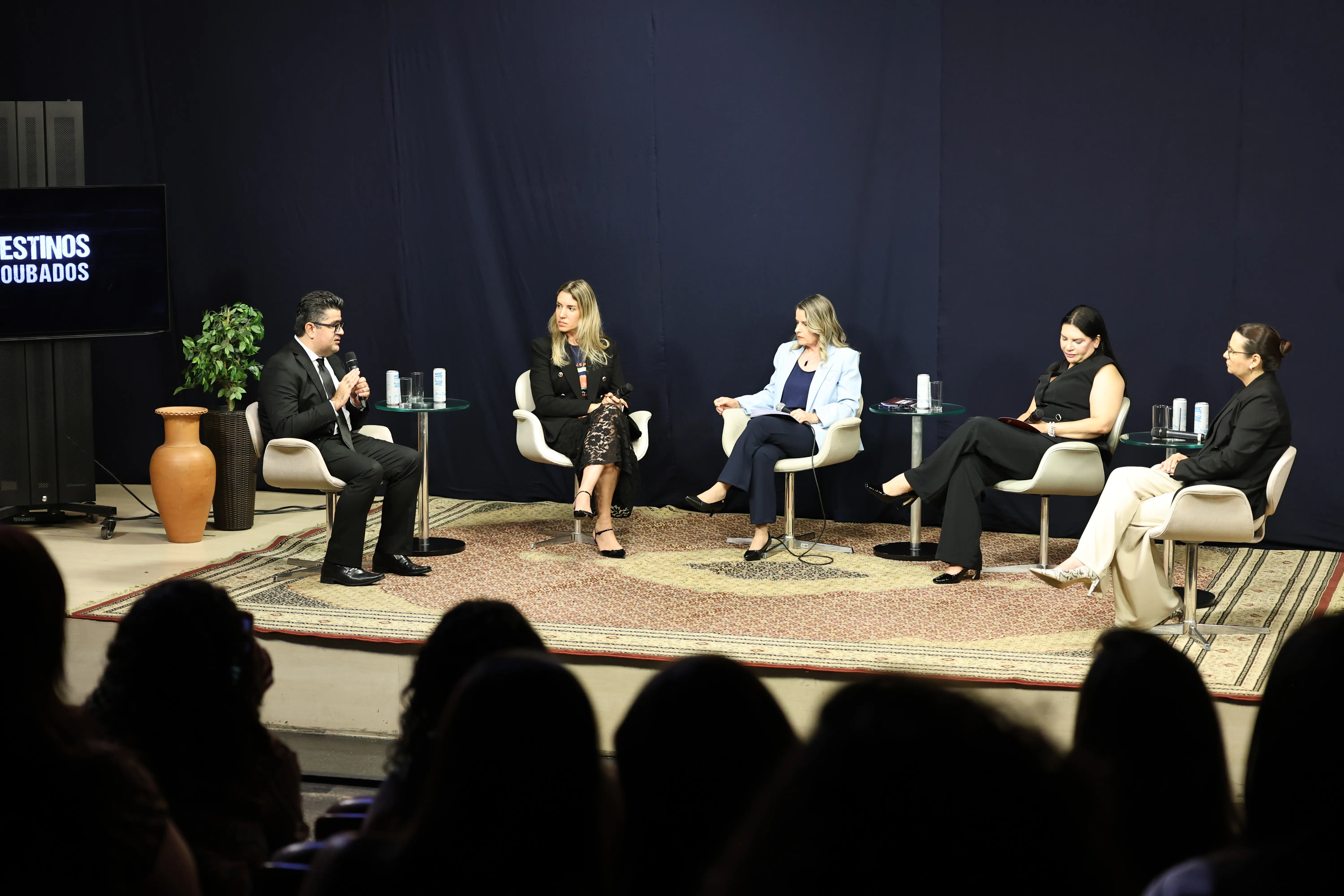 A imagem mostra cinco mulheres e um homem sentados em cadeiras brancas num palco. Todos vestem roupas formais e têm pele clara. O homem é o juiz Marcos Terêncio, que veste terno escuro e usa óculos de grau. 