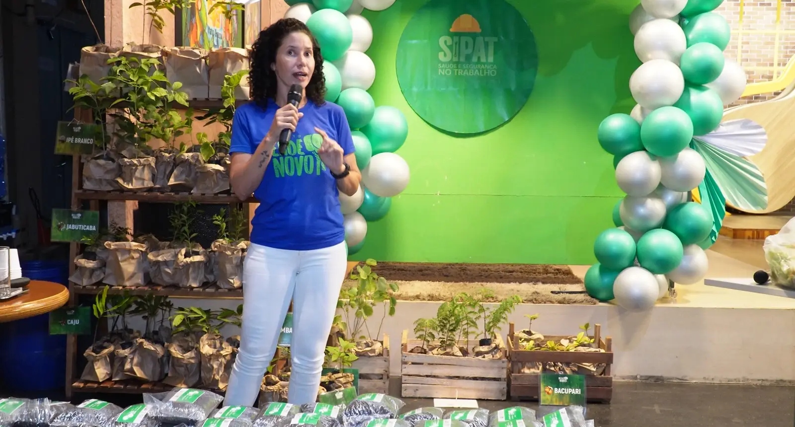Mulher de camiseta azul e calça branca fala ao microfone em frente a um painel verde da SIPAT. Ao lado, estantes de madeira com mudas de plantas em sacos de papel.