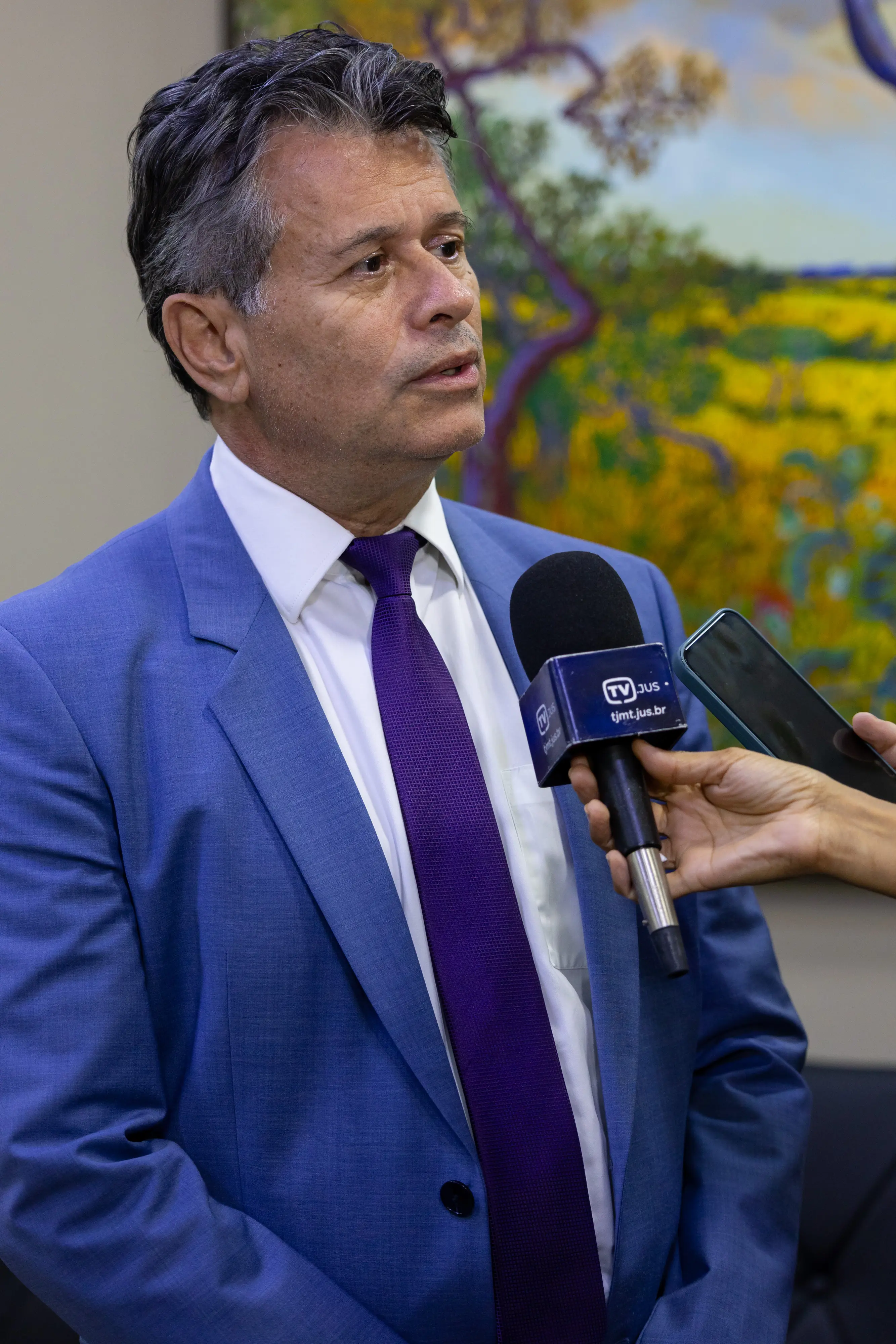 Homem de terno azul e gravata roxa sendo entrevistado. Ele olha para a direita enquanto fala em um microfone da 