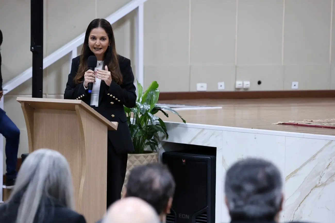 Foto horizontal que mostra a presidente da OAB Mato Grosso, Gisela Cardoso, falando ao microfone, no púlpito. Ela é uma mulher branca, de longos cabelos castanhos e lisos, usando blusa branca e blazer preto.
