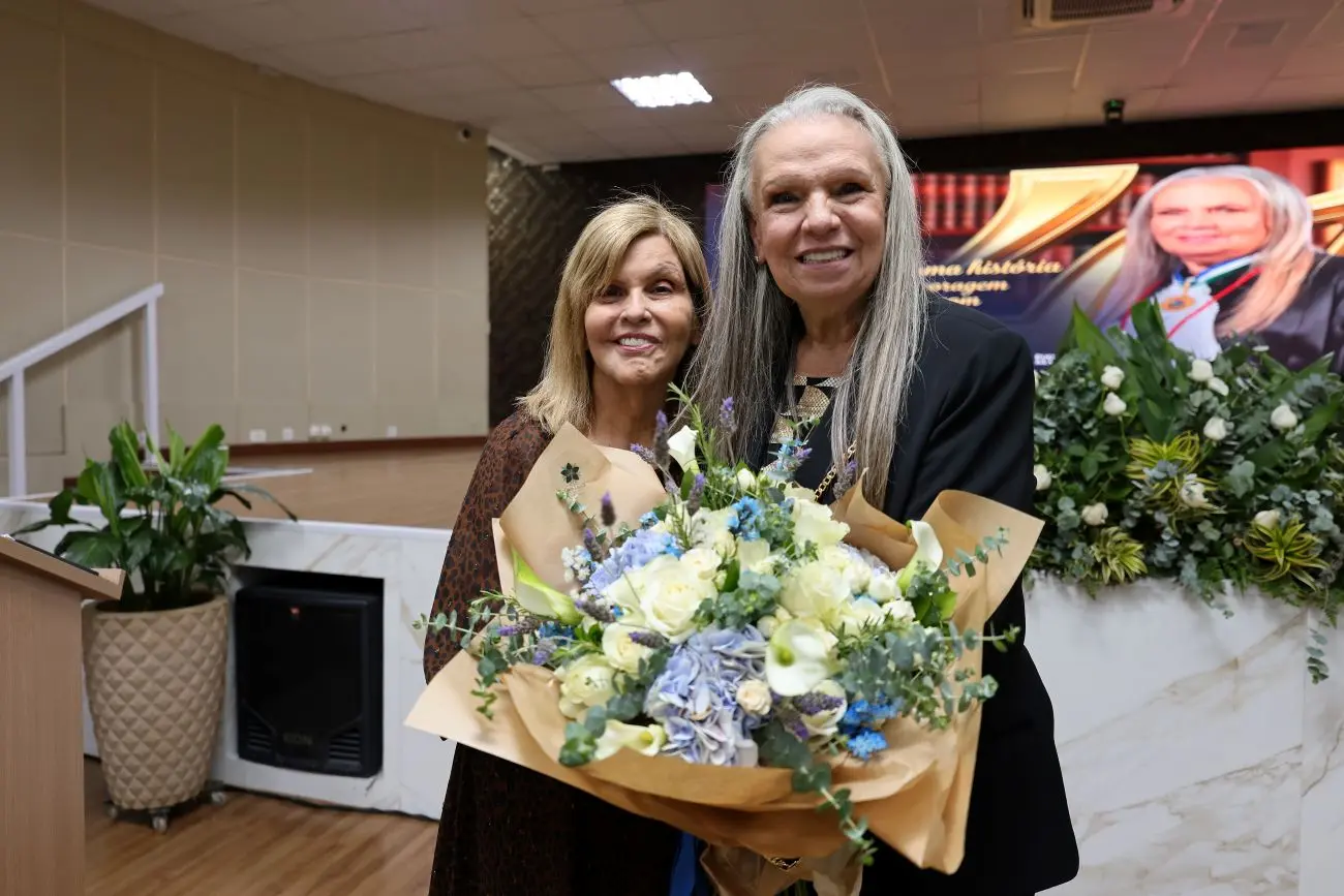 Foto horizontal que mostra as desembargadora Nilza Maria Pôssas de Carvalho e Maria Erotides Kneip abraçadas de lado, sorrindo para a foto e mostrando um buquê de flores.