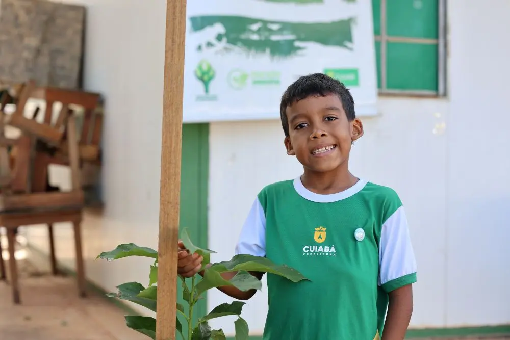 Foto horizontal que mostra o estudante Jó Abner, de oito anos, sorrindo para a foto, em pé, ao lado de uma muda de árvore plantada no pátio da escola municipal Dom Bosco. O menino tem a pele negra, olhos e cabelos castanhos, usando uniforme verde e branco.