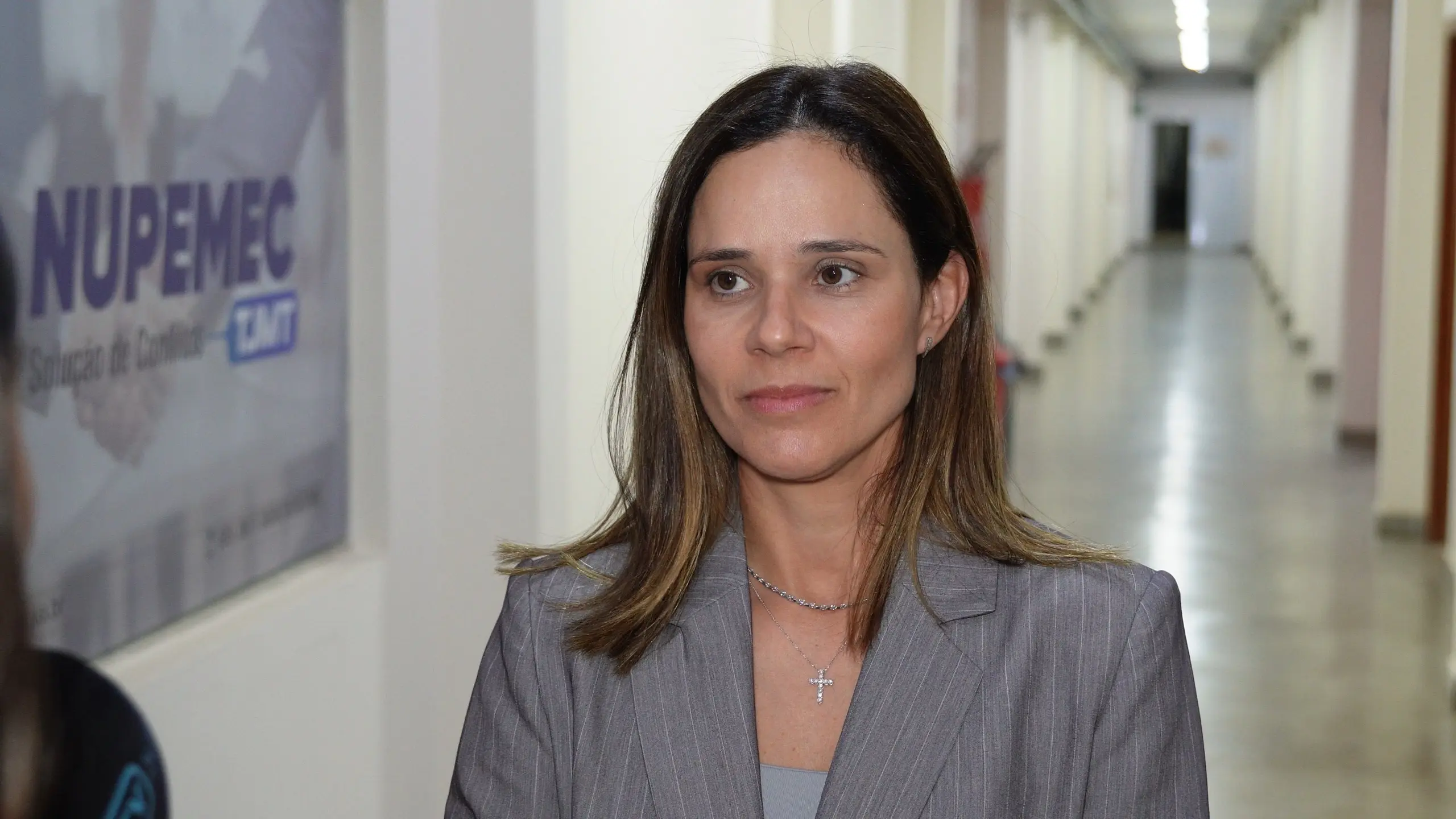 A diretora do Daje, Shusiene Machado, é uma mulher de pele clara, cabelos castanhos lisos na altura dos ombros, veste blazer cinza sobre blusa da mesma tonalidade e usa colar com pingente em formato de cruz. Ela está em um corredor, ao fundo há uma placa de identificação do Nupemec.