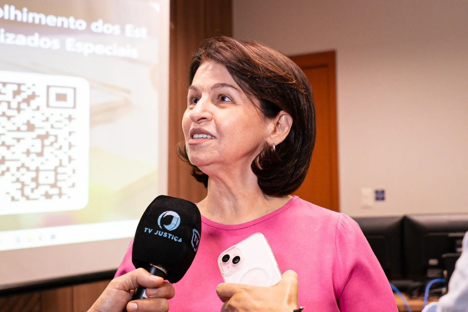 Foto horizontal que mostra a juíza Valdeci Moraes Siqueira falando ao microfone da TV Justiça, durante entrevista. Ela é uma mulher branca, de cabelos castanhos, lisos, na altura dos ombros, usando camiseta rosa. atrás dela, há um telão com um QR code projetado.