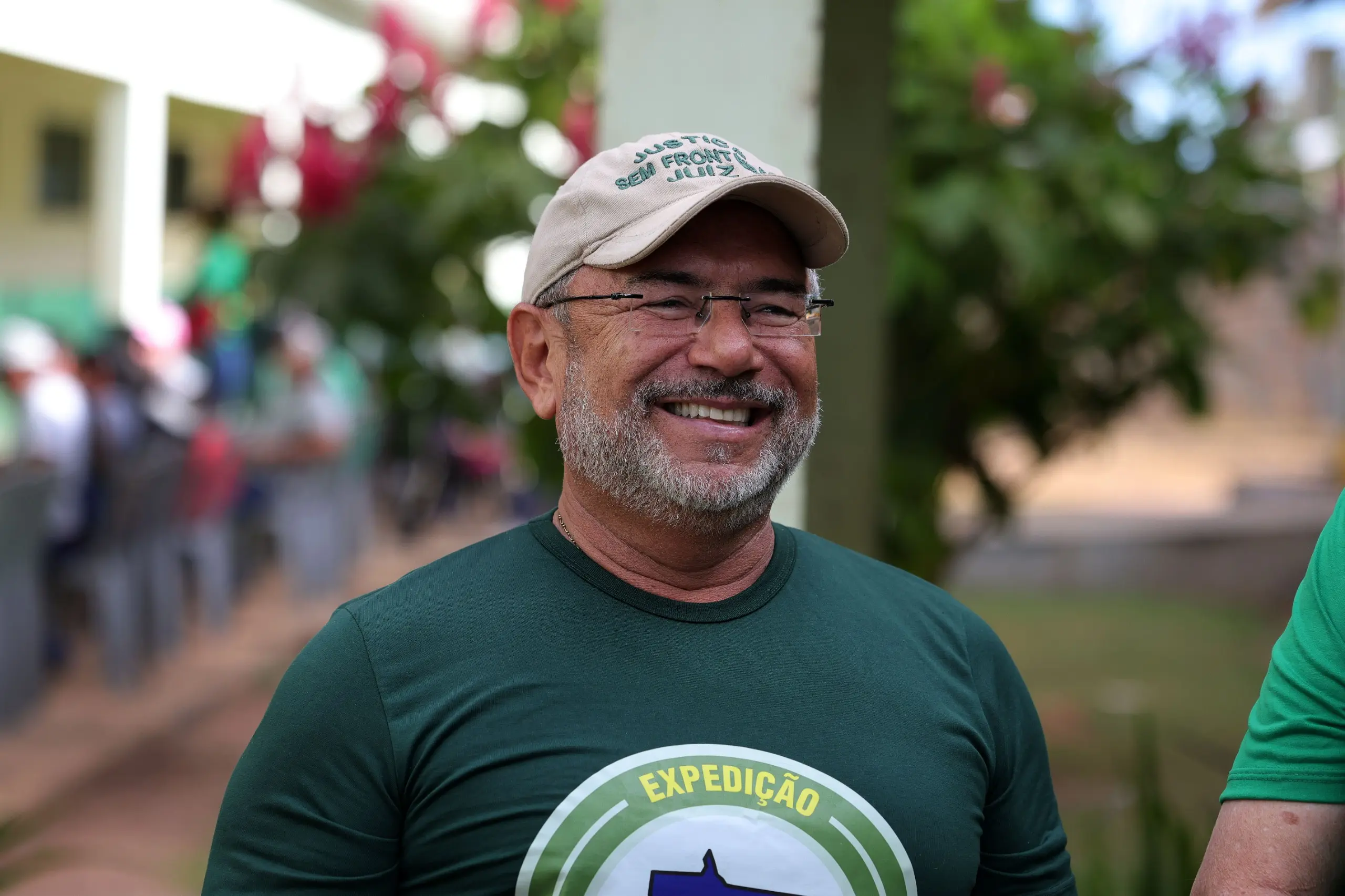 Homem sorridente de barba grisalha, óculos de grau e boné bege escrito 
