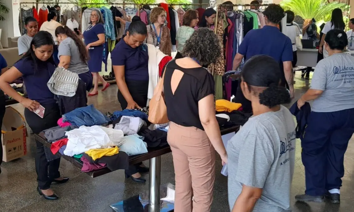 Mulheres percorrem araras e mesas repletas de roupas em salão amplo e iluminado. Ao fundo, plantas decoram o espaço movimentado.