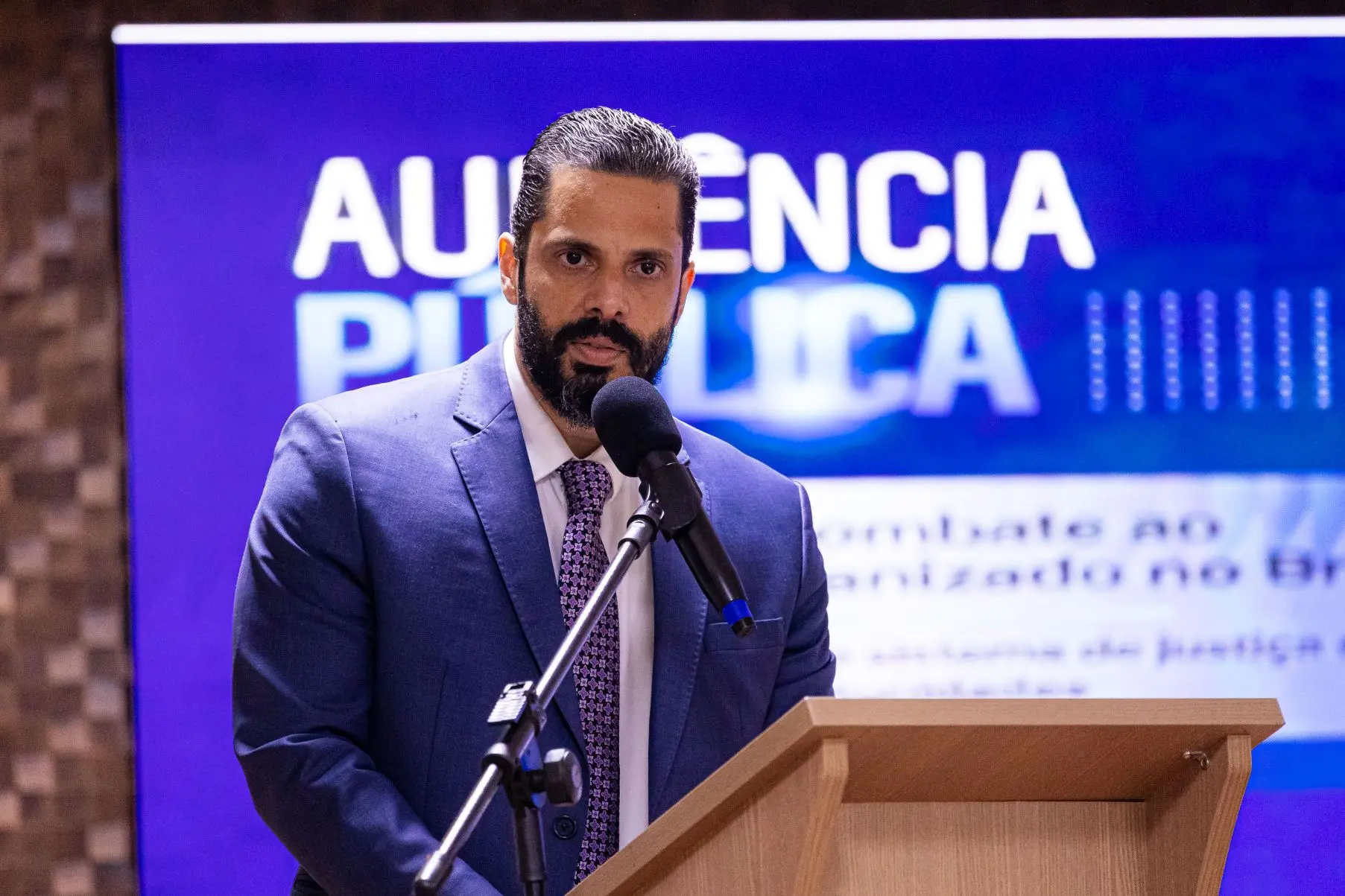 Foto horizontal que mostra o delegado de polícia Valter de Melo Fonseca Júnior do busto pra cima, falando ao microfone. Ele é um homem branco, alto, de cabelo e barba pretos, usando camisa branca, gravata azul estampada e terno azul. Ao fundo, um telão com as informações da audiência pública.