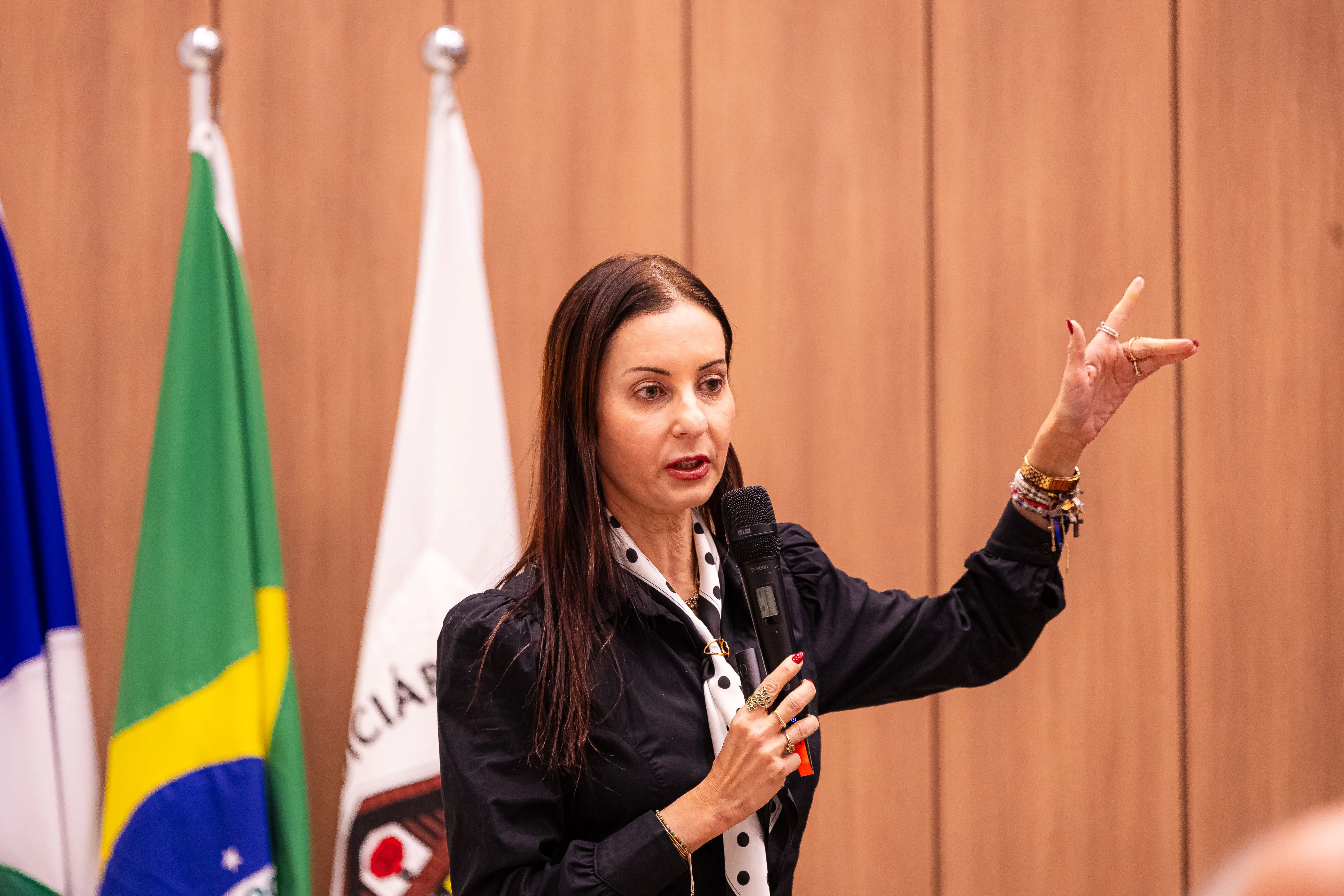 Mulher de cabelos longos e escuros, vestindo blusa preta com detalhes em branco, gesticula com a mão esquerda enquanto fala ao microfone em um evento.