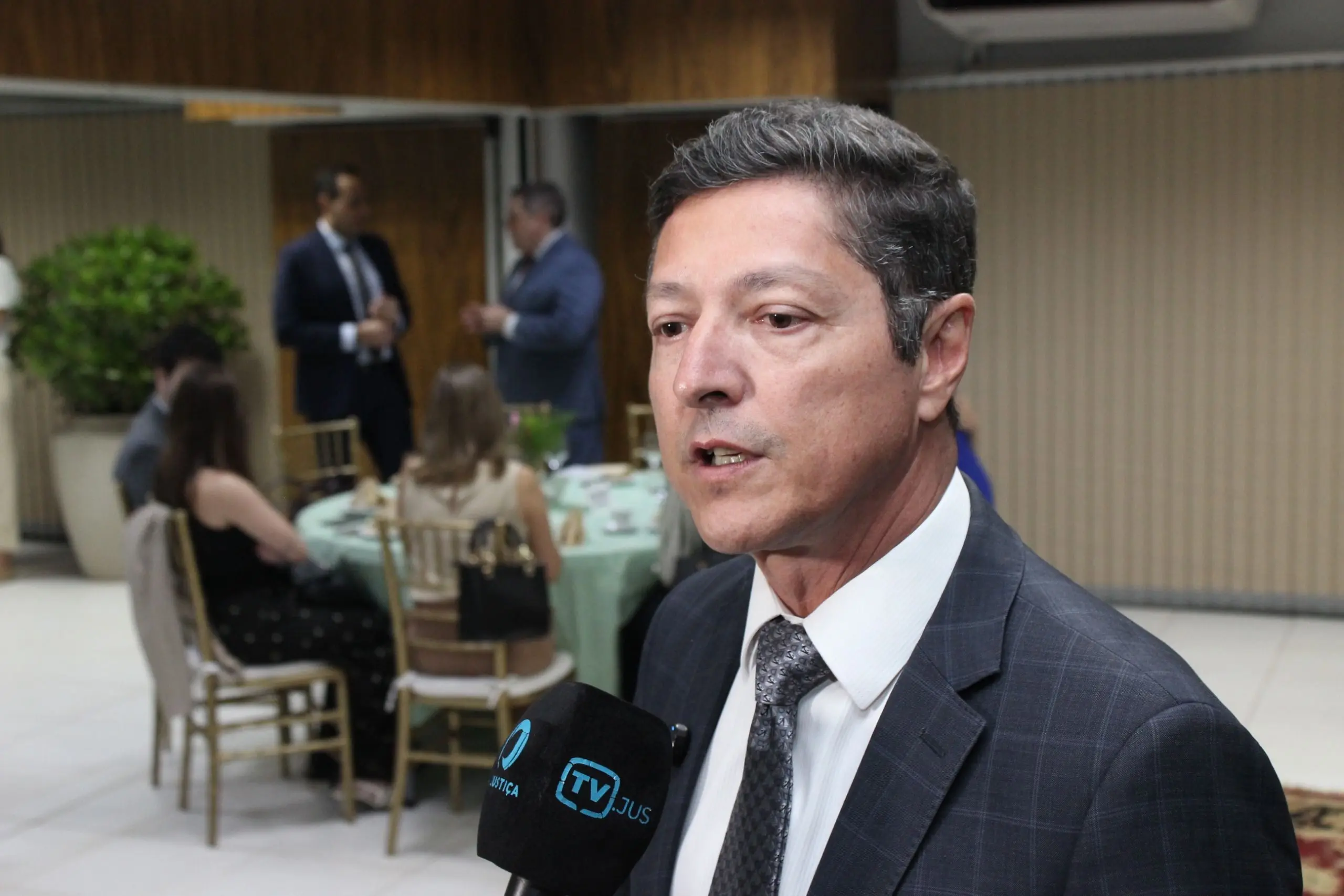 Pessoa em traje social concede entrevista em ambiente interno, falando diante de um microfone com logotipo. Ao fundo, há mesas com pessoas sentadas e outras em pé, além de parede com revestimento de madeira e iluminação suave, indicando evento institucional.