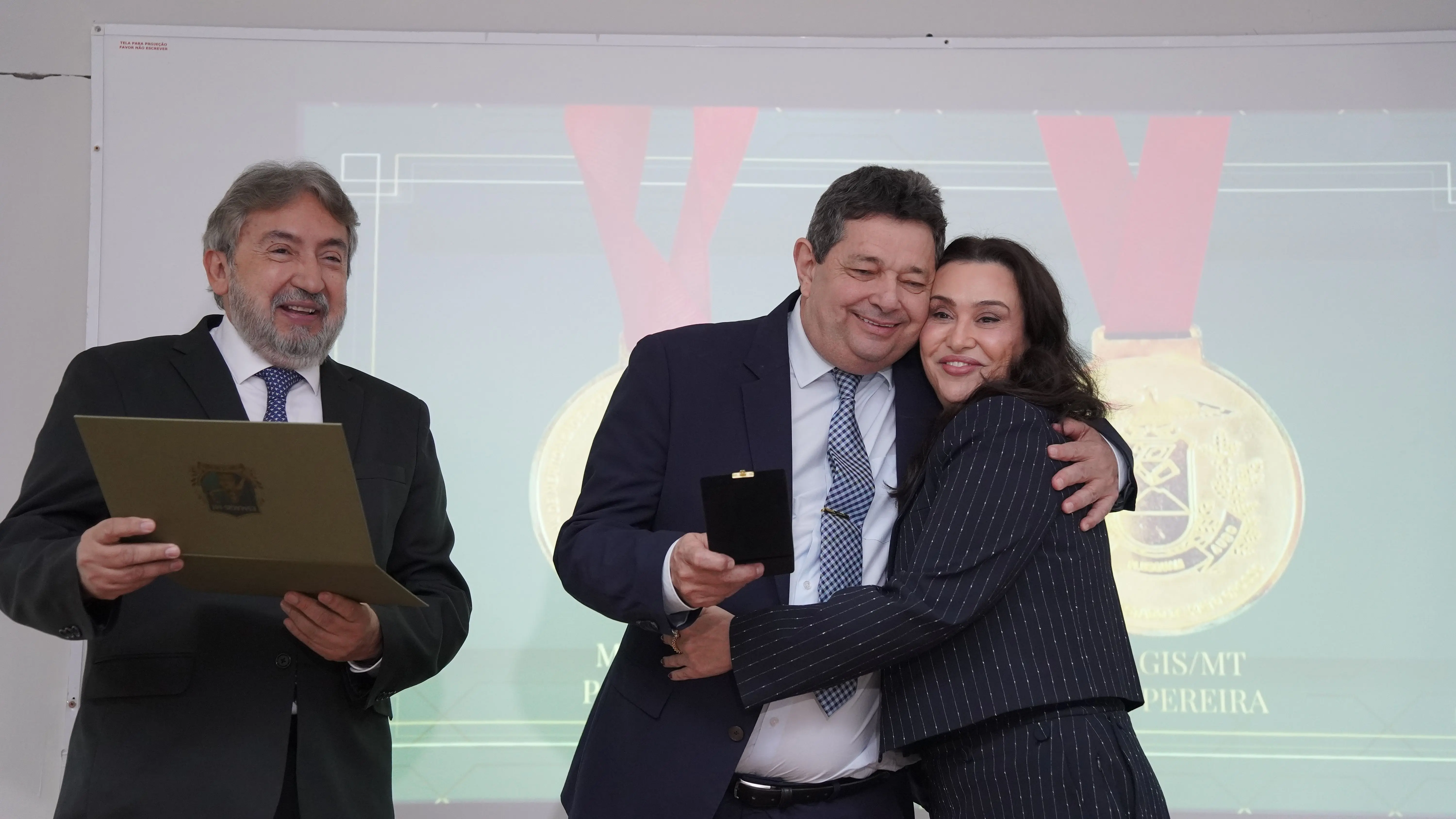 Homem de terno abraça sorridente uma mulher de terninho listrado enquanto segura uma caixinha de medalha. À esquerda, outro homem segura um diploma. Ao fundo, a projeção de uma grande medalha de ouro.