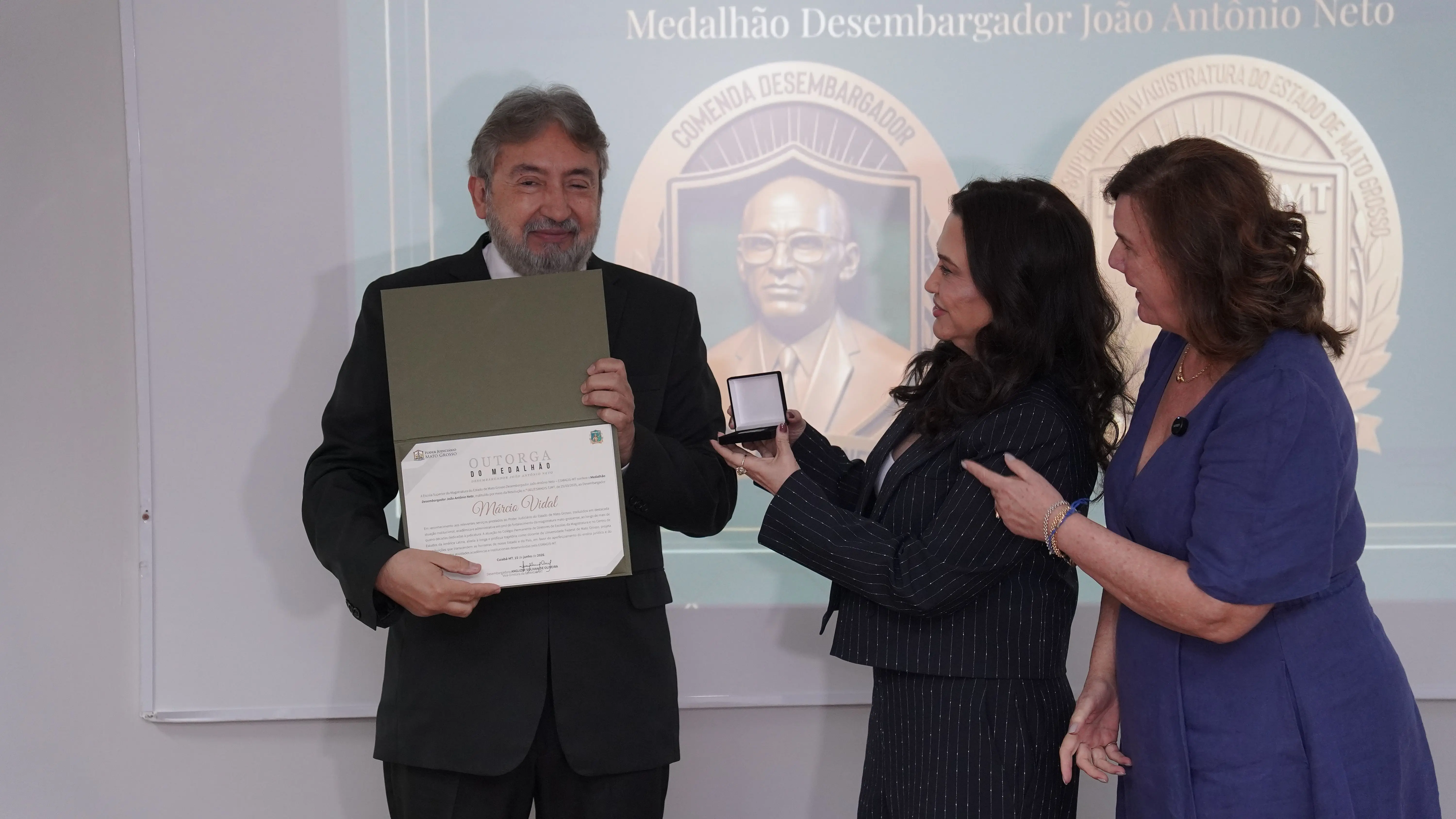 Homem de terno preto segura um diploma aberto enquanto duas mulheres, uma de terninho listrado e outra de vestido azul, entregam uma caixinha de medalha. Ao fundo, projeção com brasões institucionais.