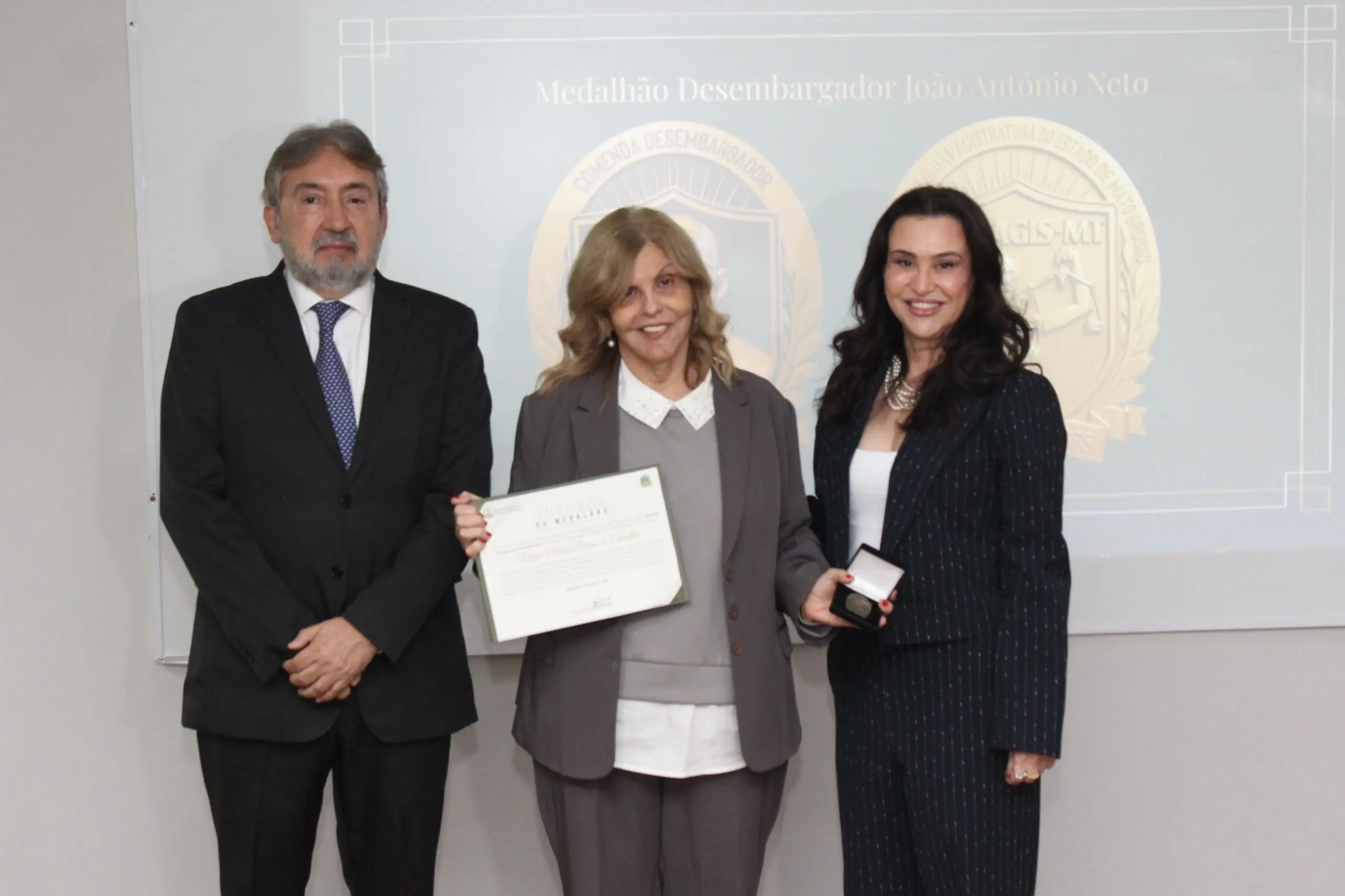 Mulher ao centro exibe diploma ao lado de um homem de terno e de outra mulher que segura uma caixinha de medalha. Ao fundo, a projeção indica 