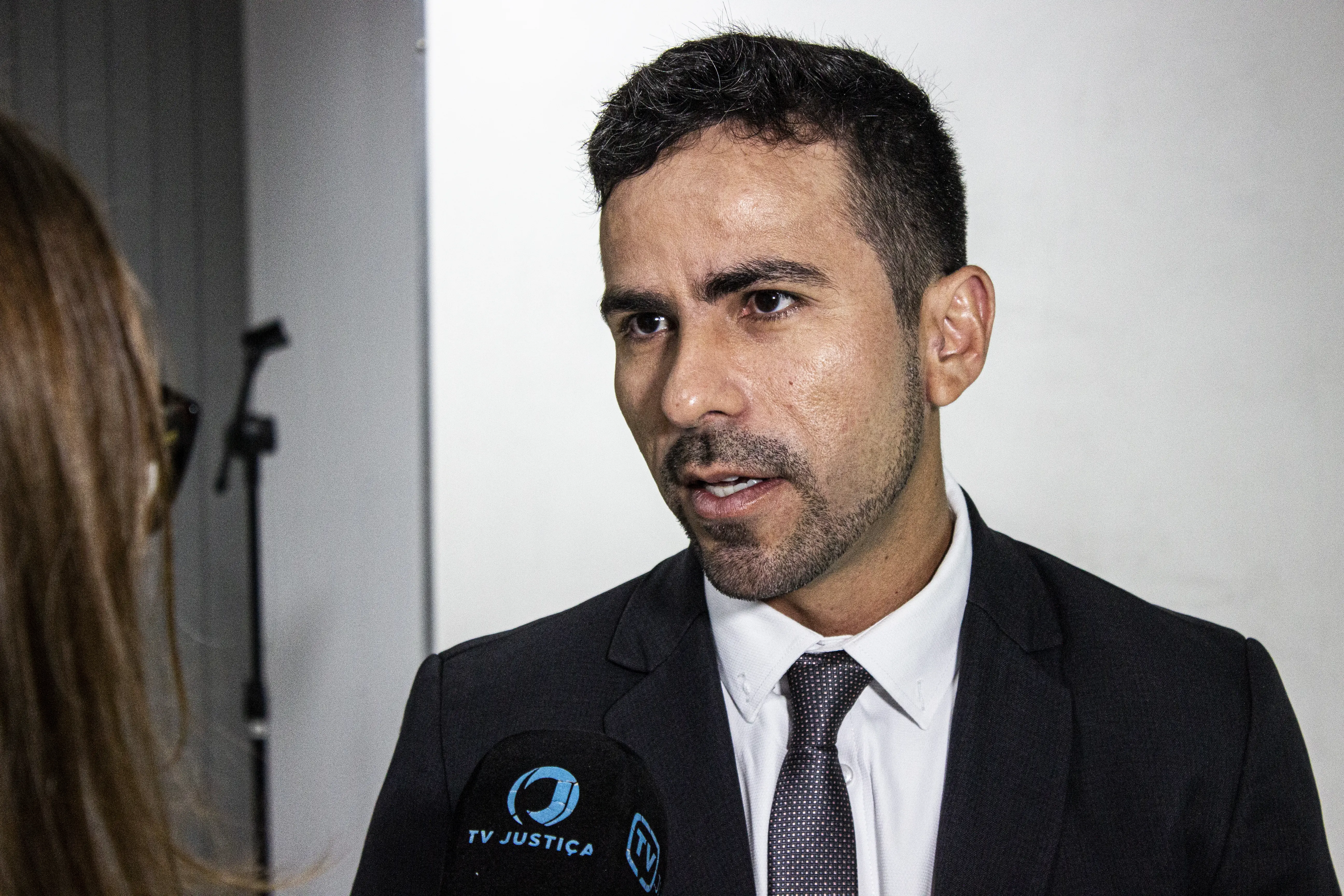 Homem de terno escuro, camisa branca e gravata é entrevistado por pessoa parcialmente visível à esquerda. Ao centro da imagem, aparece um microfone voltado para ele.