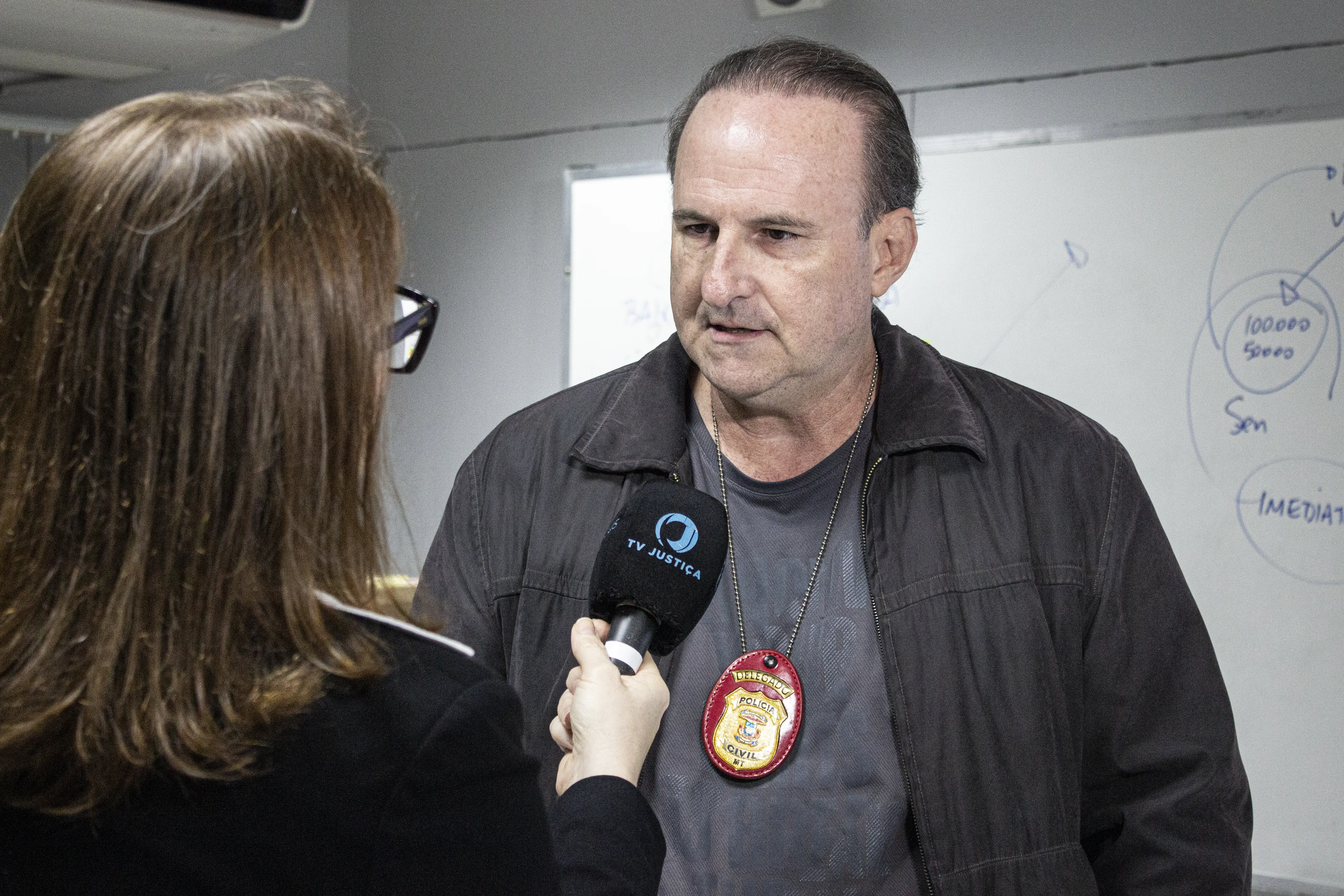 Homem usa jaqueta escura e distintivo pendurado no pescoço enquanto concede entrevista. Uma pessoa segura microfone voltado para ele diante de quadro branco.