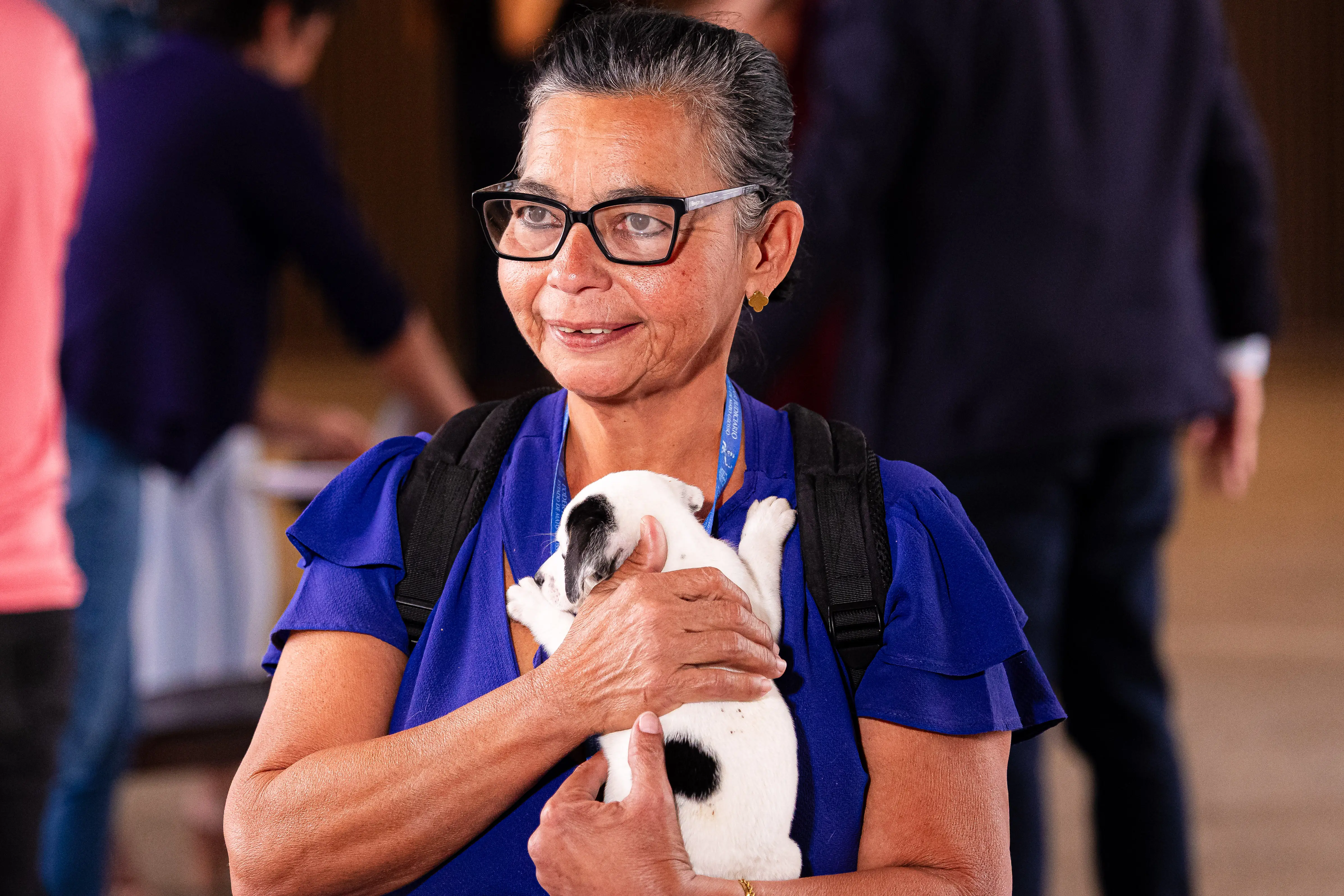 A servidora Michela Aparecida Neves Pereira segura nos braços um filhote de cachorro branco com manchas pretas recém-adotado durante a feira de adoção de animais. Ao fundo, participantes circulam pelo local do evento.