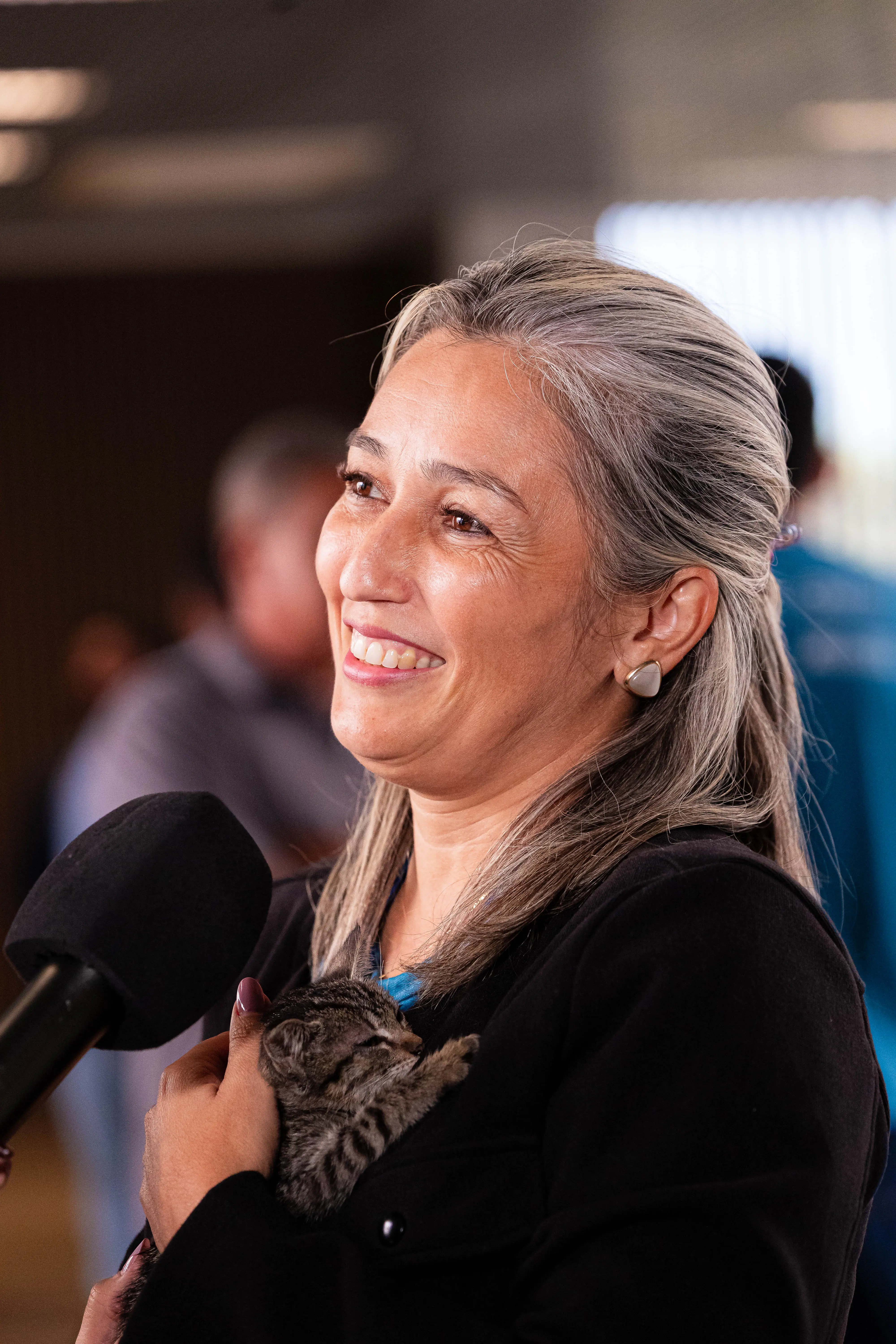 A servidora da Corregedoria Gracyelly Arruda segura nos braços um filhote de gato tigrado durante a feira de adoção de animais, enquanto concede entrevista. Ao fundo, participantes circulam pelo local do evento.