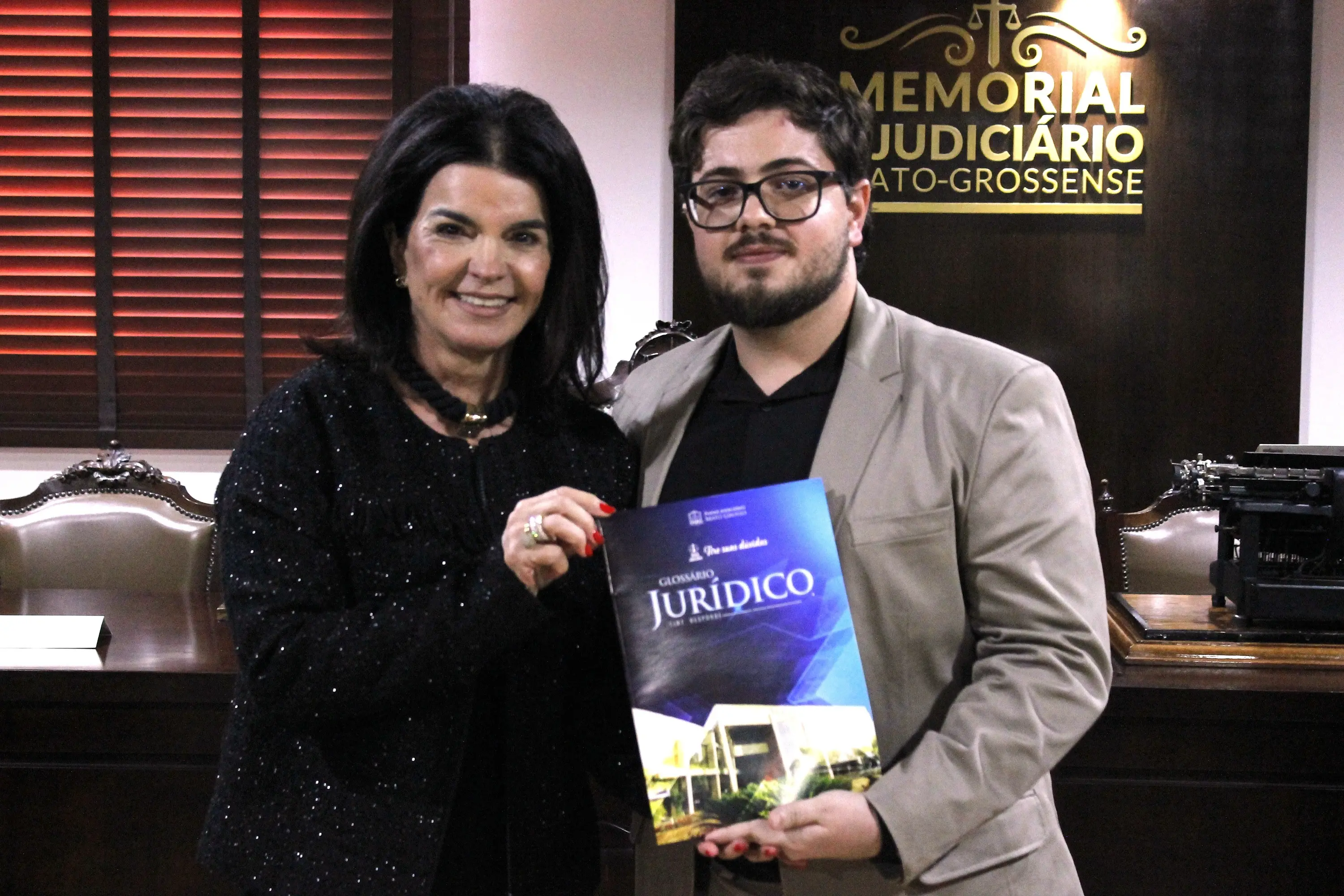 Mulher de cabelos pretos e homem jovem de óculos e blazer bege seguram um Glossário Jurídico no Memorial do Judiciário Mato-Grossense.