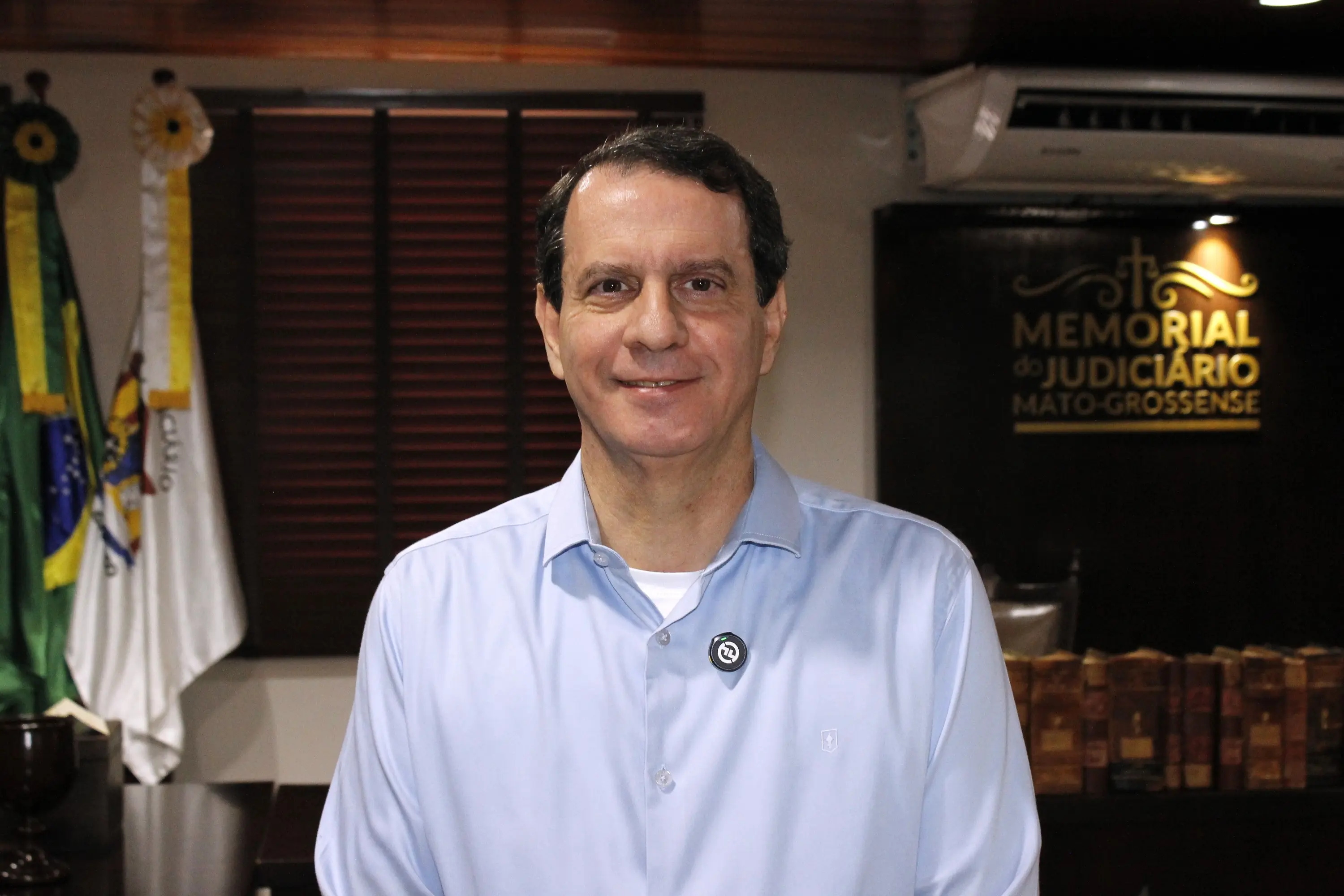 Homem de cabelos escuros, camisa azul-claro sorri para a câmera. Ao fundo, bandeiras e o painel do Memorial do Judiciário Mato-Grossense.