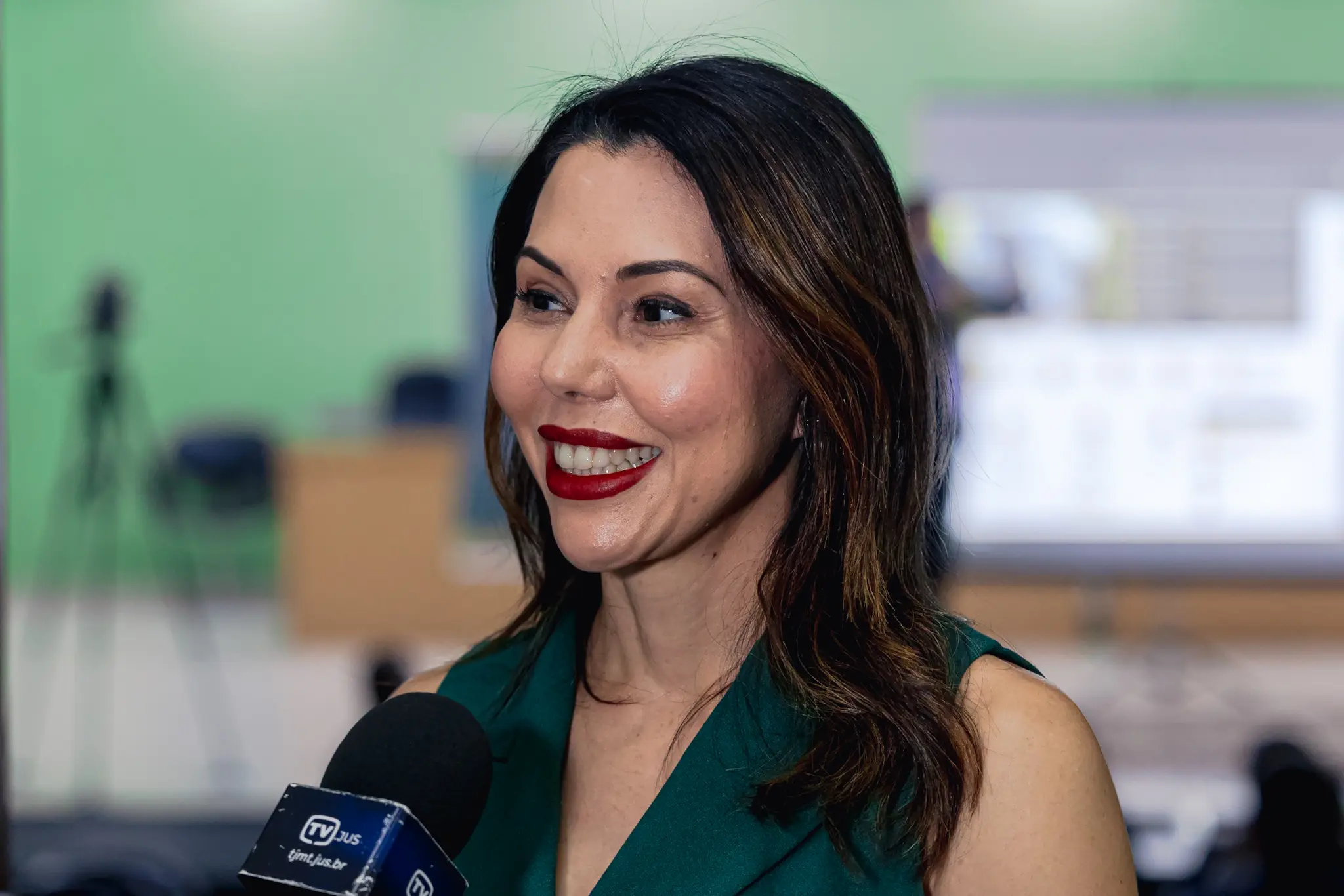 Mulher de cabelos escuros e batom vermelho sorri ao conceder entrevista. Ela veste blusa verde-escura sem mangas. Um microfone da 