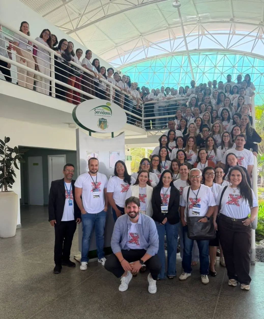 Grande grupo de pessoas reunido em um saguão moderno com mezanino e teto arqueado. Muitos vestem camisetas brancas de campanha. Um homem de camisa clara e tênis branco agacha-se à frente de todos.