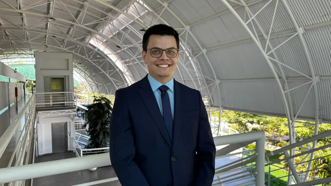 Foto em plano médio do juiz Lessandro sorrindo, vestindo terno azul marinho, camisa azul claro, gravata azul marinho e óculos. Ele é branco de cabelos castanhos, está em uma passarela com estrutura metálica branca, telhado em arco e treliças brancas.