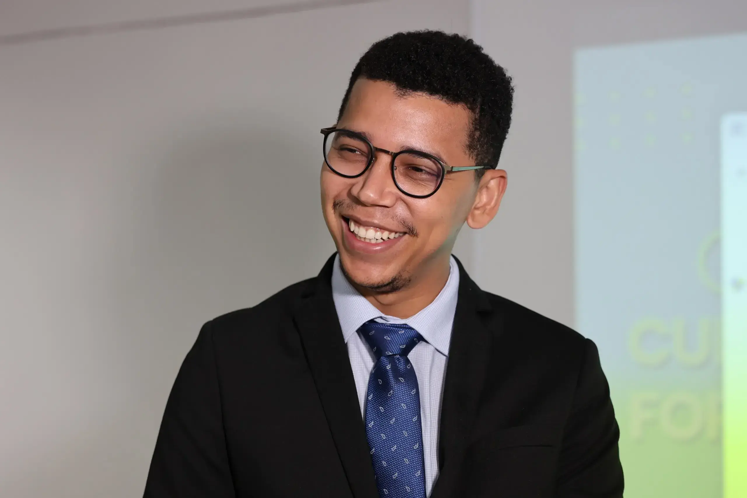 Foto horizontal do juiz Thiago dos Santos, que aparece sorridente em um plano médio, vestindo terno preto, camisa social clara e gravata azul. Ele usa óculos, possui cabelos curtos e crespos e uma barba rala bem aparada. 
