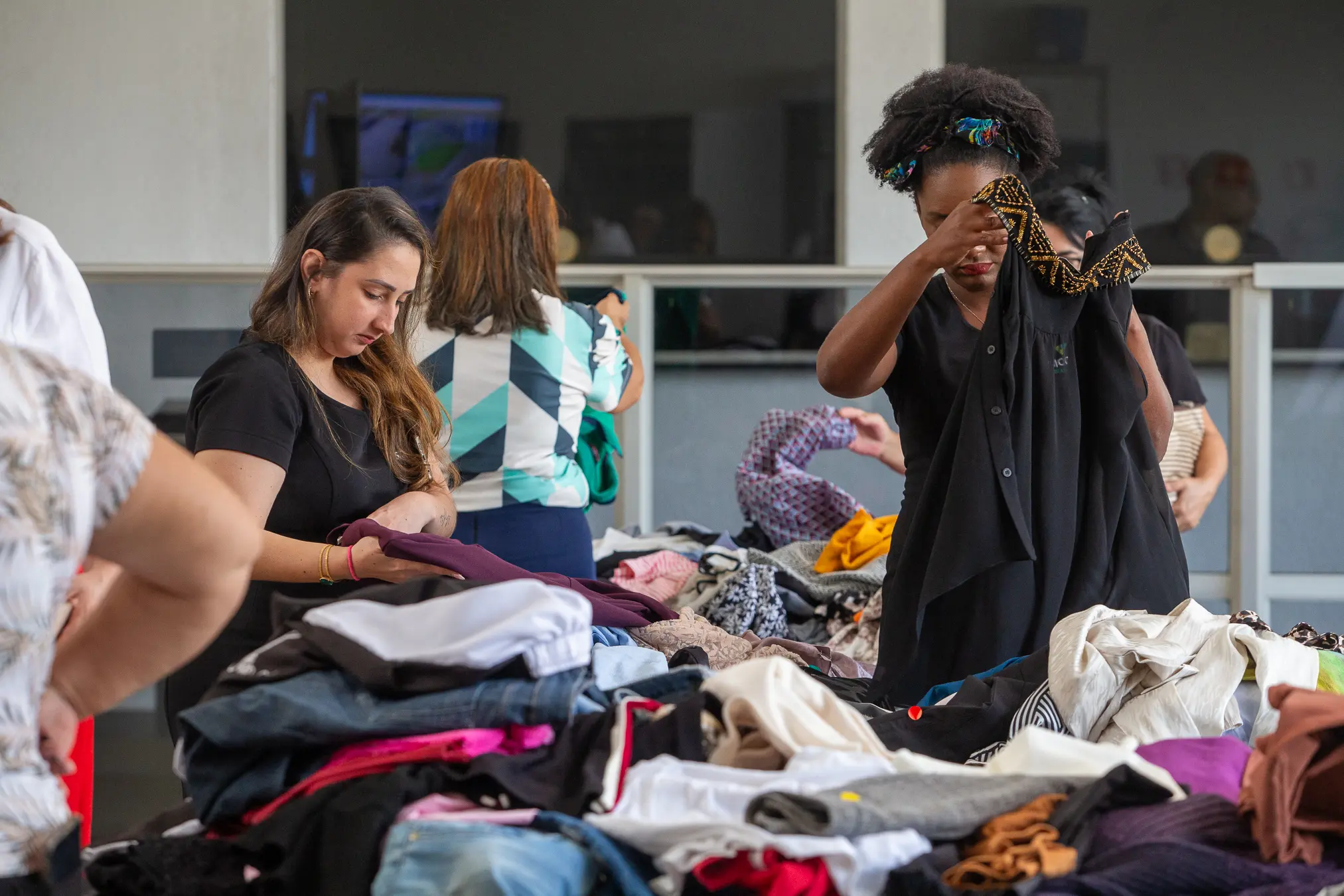 Mulheres vasculham pilhas de roupas coloridas sobre mesas. Em destaque, jovem de blusa preta e cabelos cacheados examina peça escura.