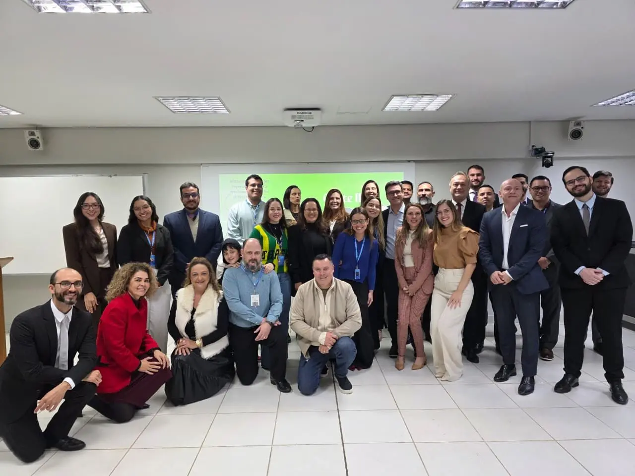 Fotografia de grupo em ambiente interno. Cerca de 30 pessoas posam para a foto em uma sala com piso claro, paredes brancas e iluminação de teto. Parte do grupo está em pé e outra parte está agachada ou sentada na frente. Os participantes usam trajes sociais e crachás. Ao fundo, há uma tela branca e 
