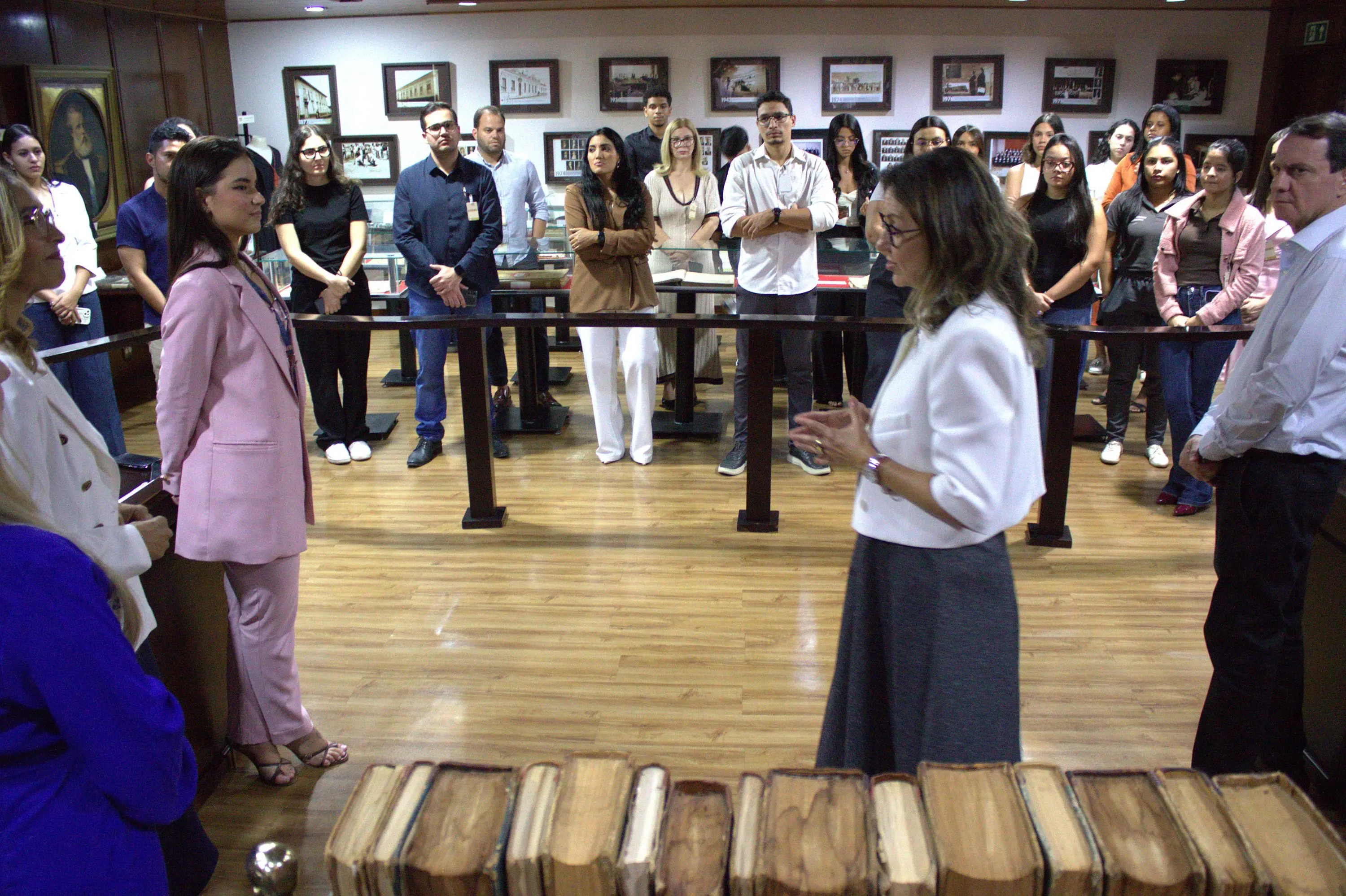 Mulher de blazer branco discursa para grupo de jovens reunidos em semicírculo em sala com vitrines e quadros históricos nas paredes.